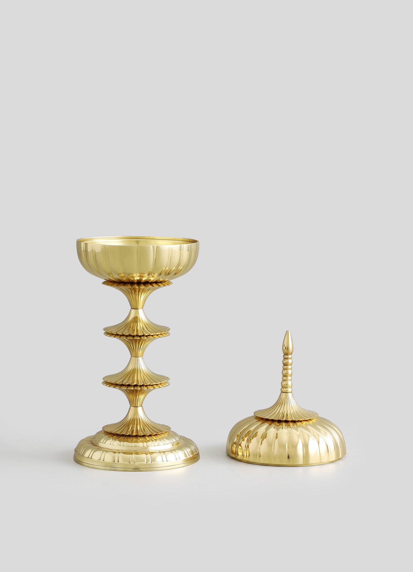 Gold-toned decorative objects with sculptural, ribbed motifs, featuring a bowl with a fluted edge and a distinctively shaped lid with a pointed finial. The items have a polished metal finish and an elegant, contemporary style.