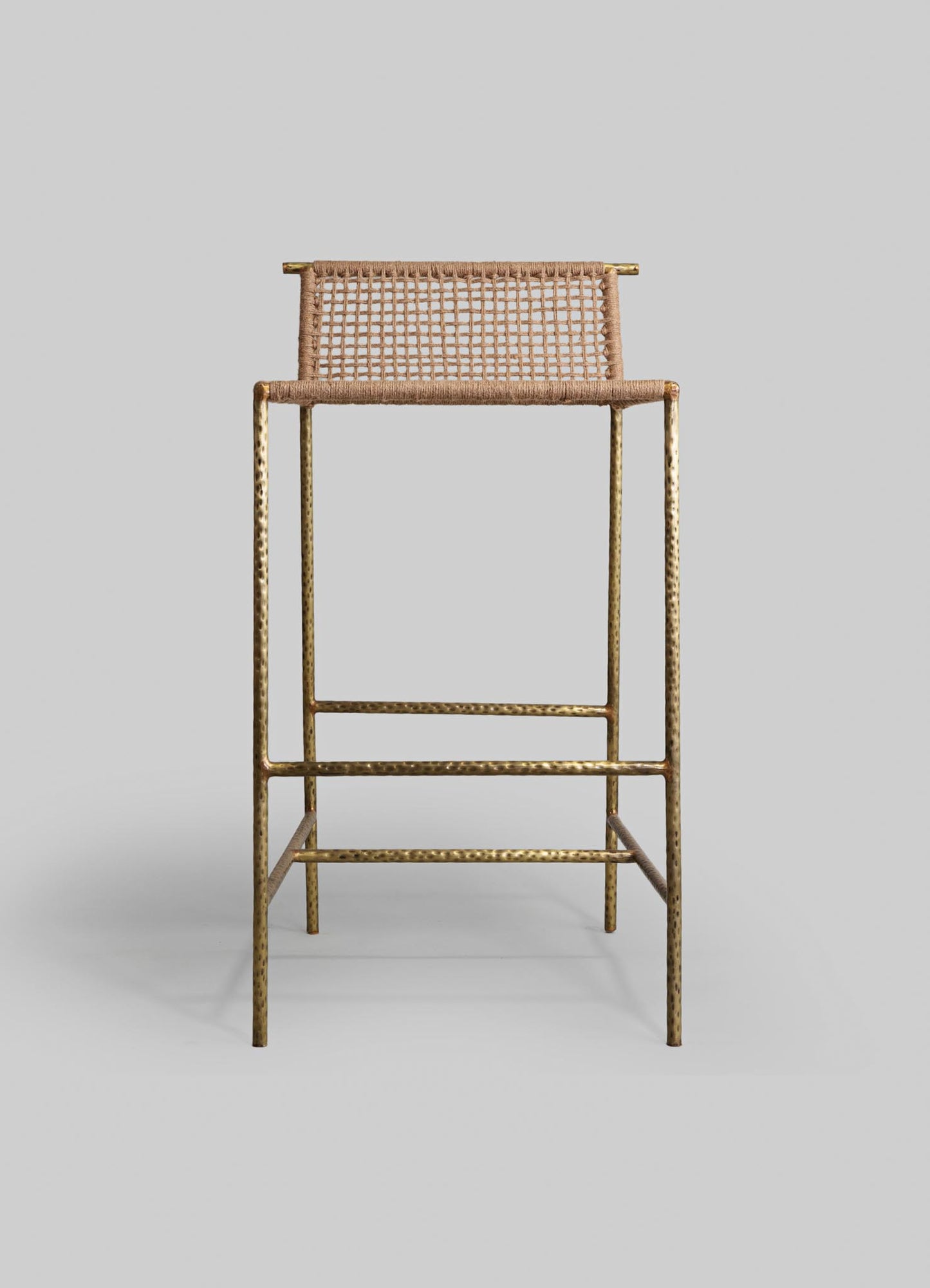 A tall stool with a brass hammered finish metal frame and a woven rattan or cane seat and backrest, featuring a natural beige tone. The design combines contemporary and rustic elements with a minimalist aesthetic.