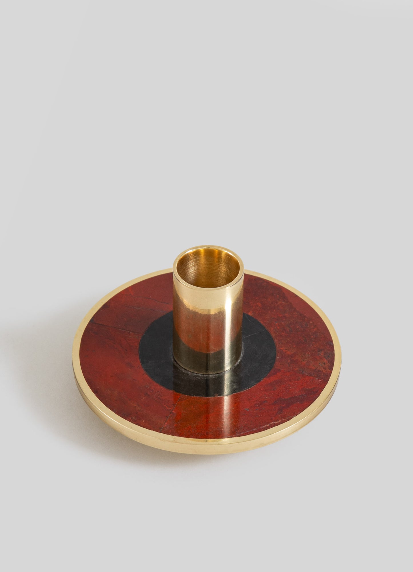 A decorative candle holder featuring a circular base with a red and black marble pattern and a brass outer edge, with a cylindrical brass holder in the center.