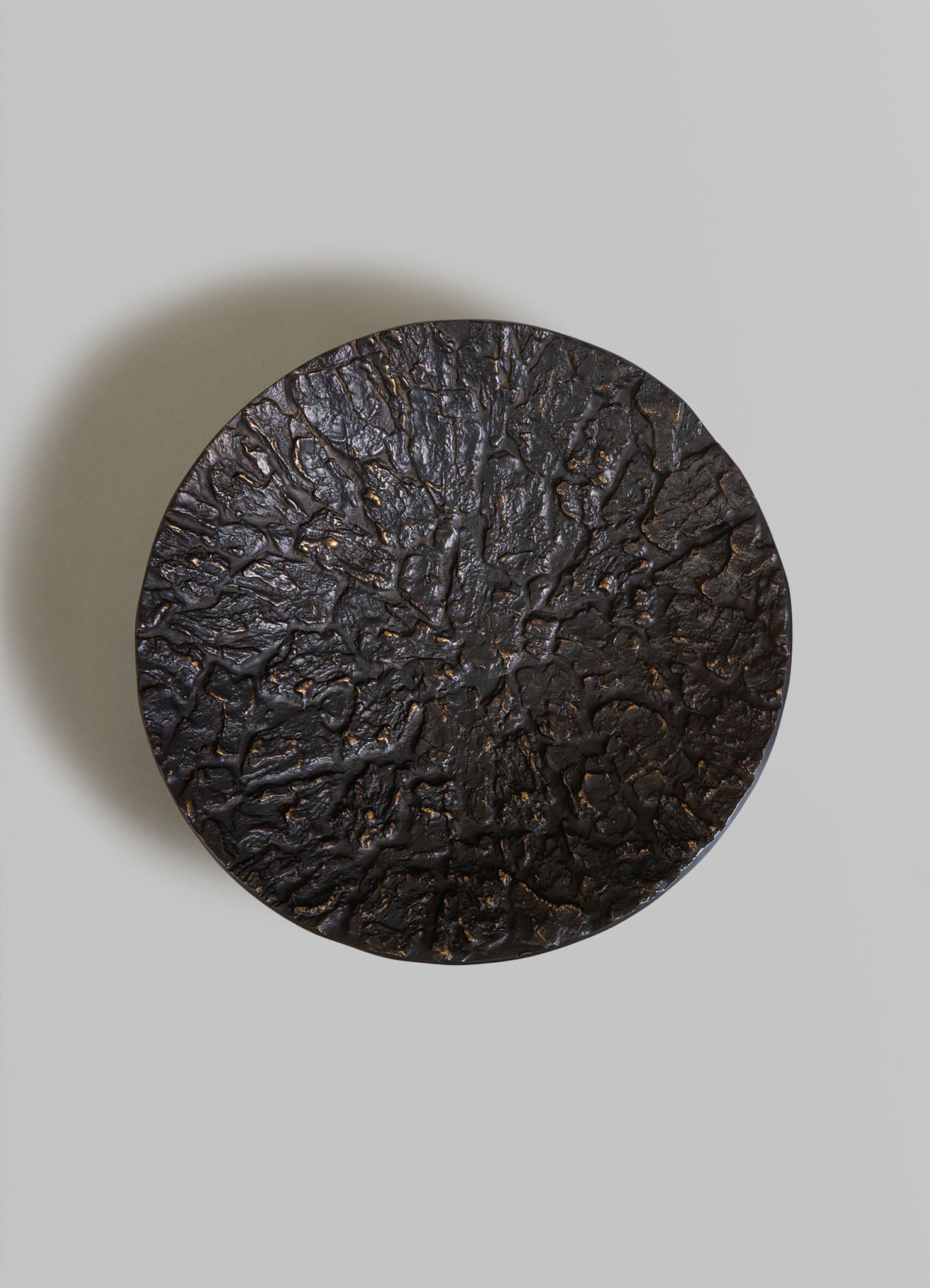 A round, textured black tray with an uneven, cracked surface resembling volcanic or natural stone, placed on a neutral background. The tray has an organic, rustic style with a dark matte finish.