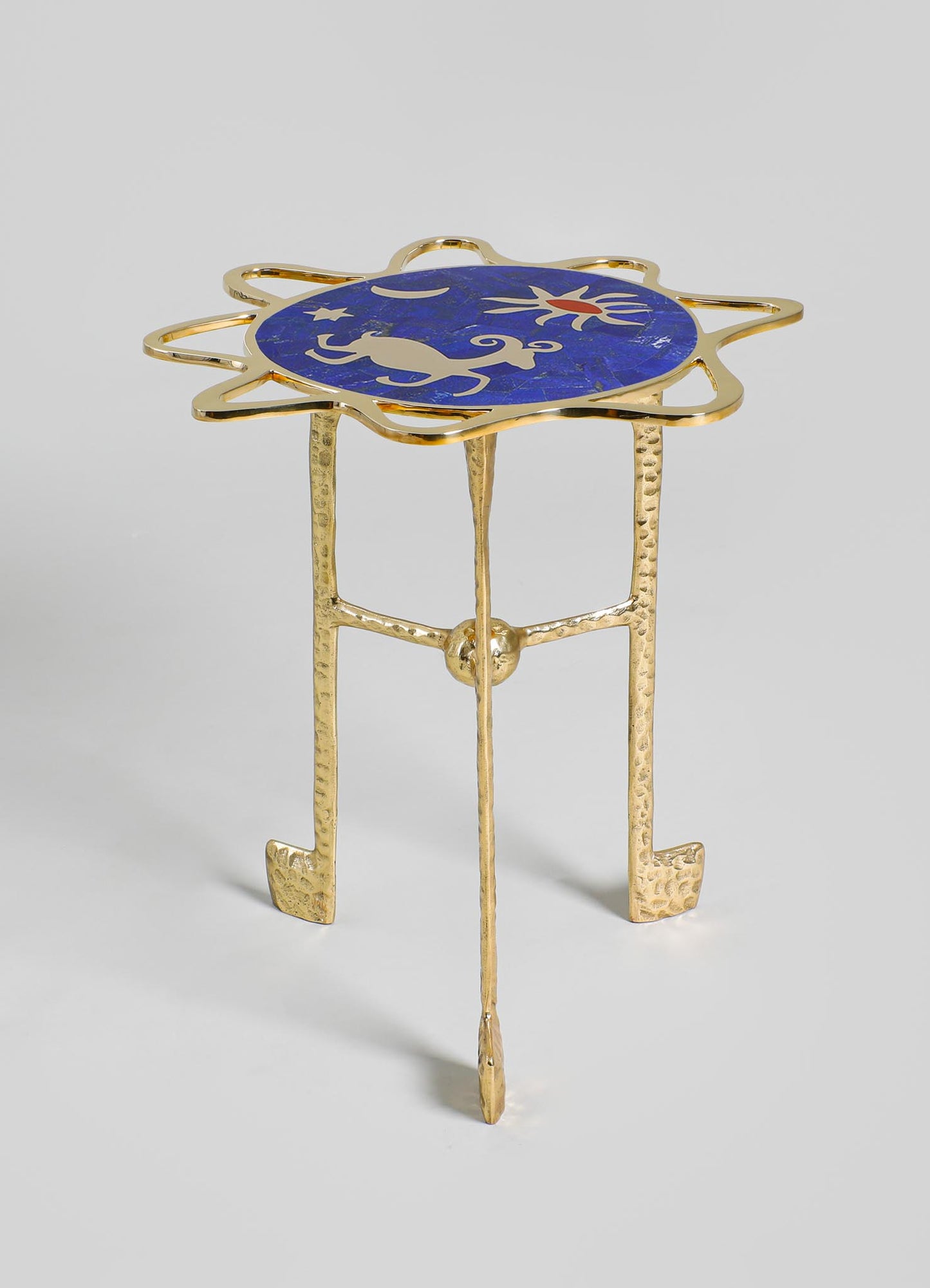 A decorative side table with a gold hammered metal frame and three legs, featuring a round tabletop with a blue enamel surface adorned with a celestial motif of stars, a crescent moon, and a sun in red and white.