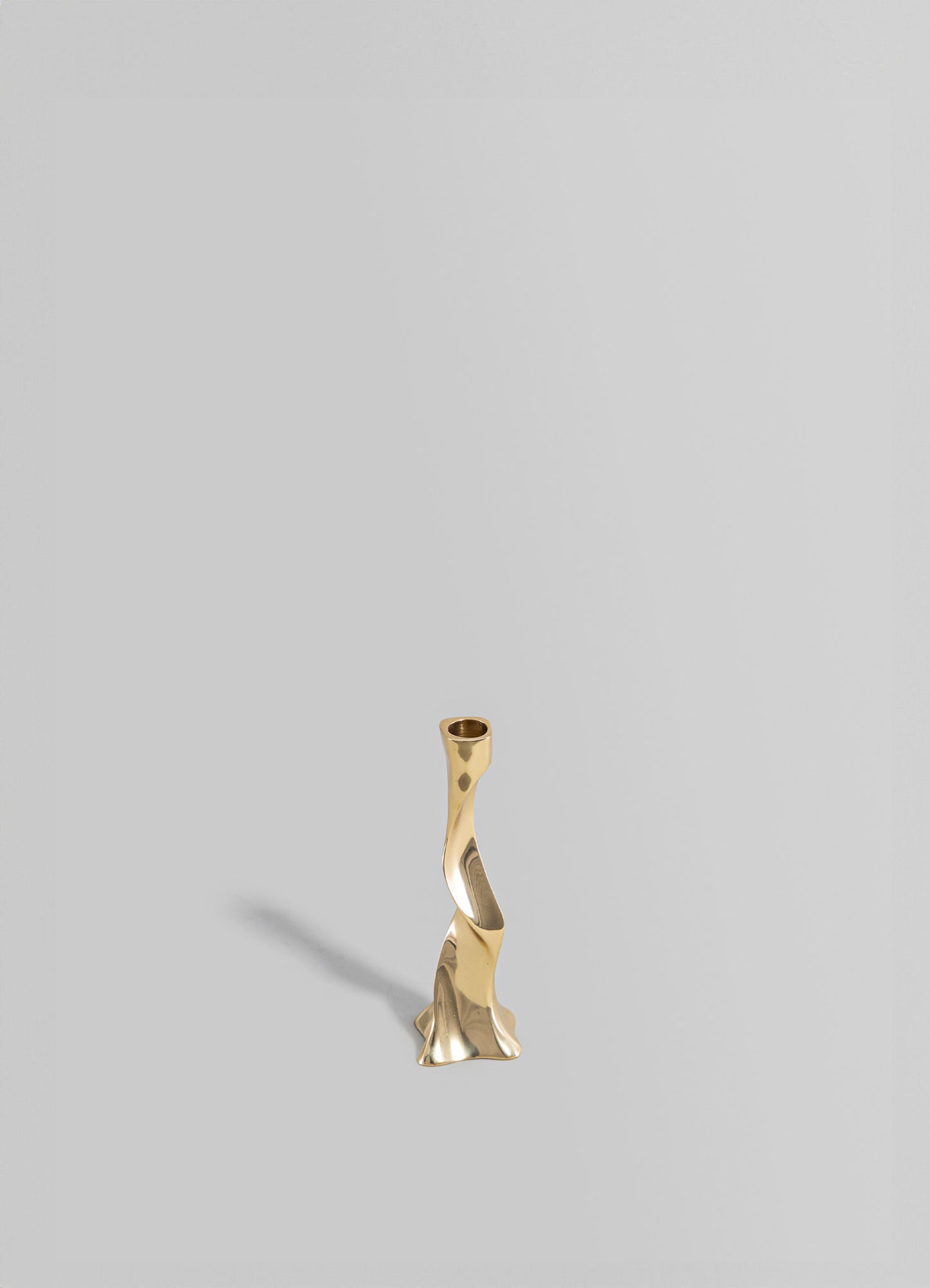 A modern sculptural brass candlestick with a twisted, organic shape and a polished gold finish, set against a plain, light gray background.