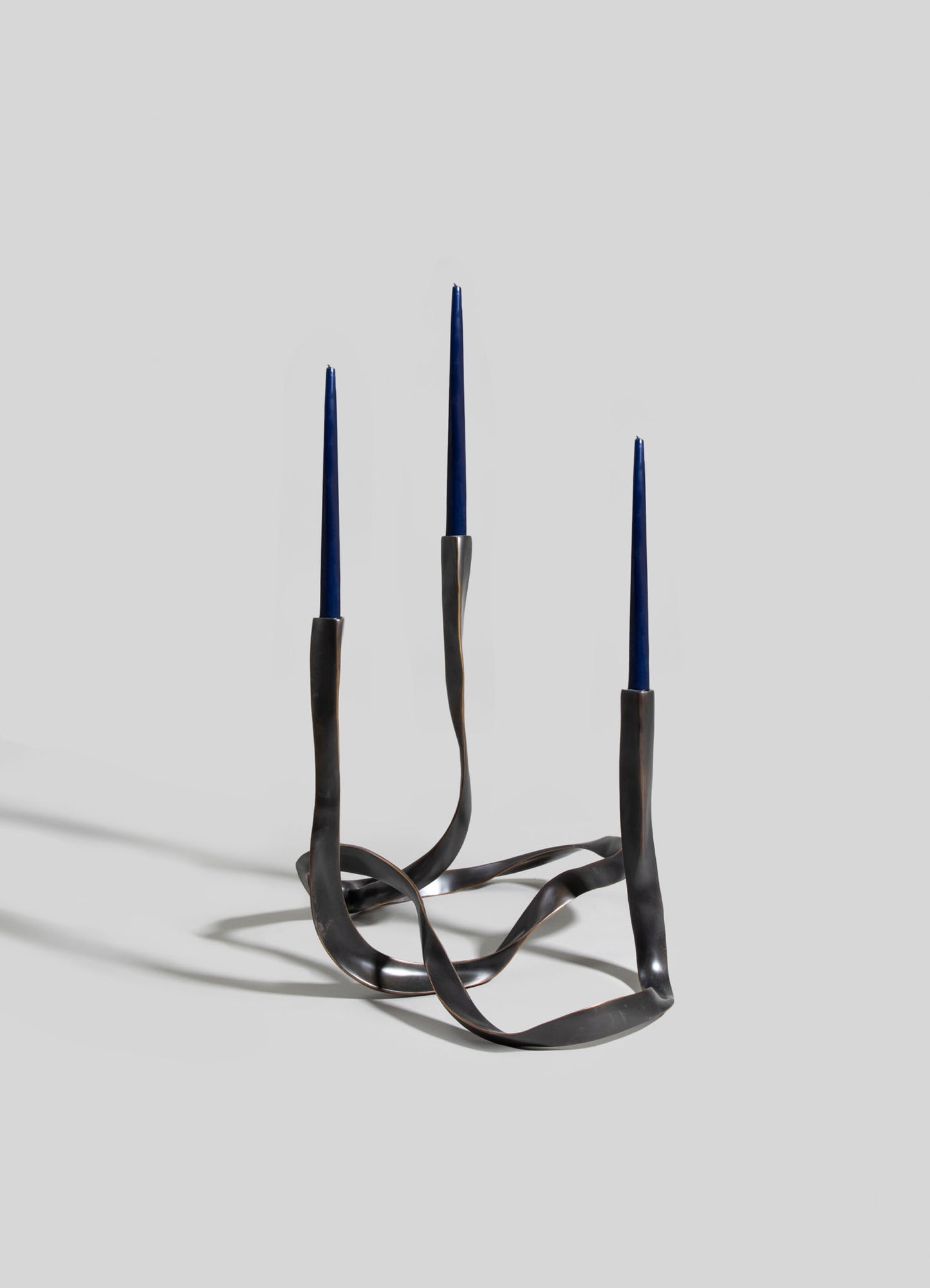 A sculptural candle holder with a twisted metal base in a dark, matte finish, holding three tall, slender blue candles. The design features organic, flowing lines and an abstract, contemporary style.