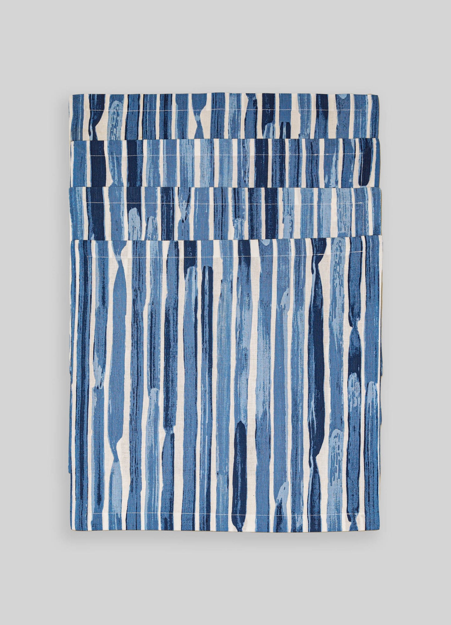 A set of fabric napkins with a blue and white striped pattern, featuring various shades of blue and irregular, hand-painted style stripes. The textiles have a contemporary, artisanal aesthetic.