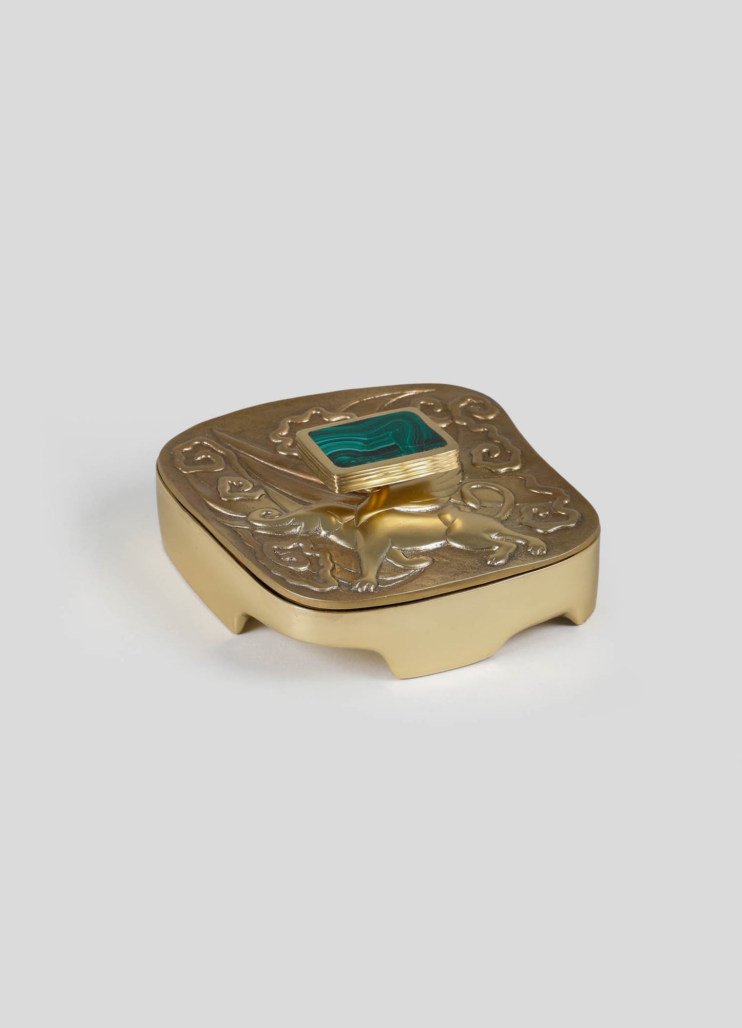A decorative jewelry box with a gold-toned finish, featuring ornate embossed motifs on the lid and a square turquoise stone in a raised setting. The box has curved legs and a vintage-inspired design.