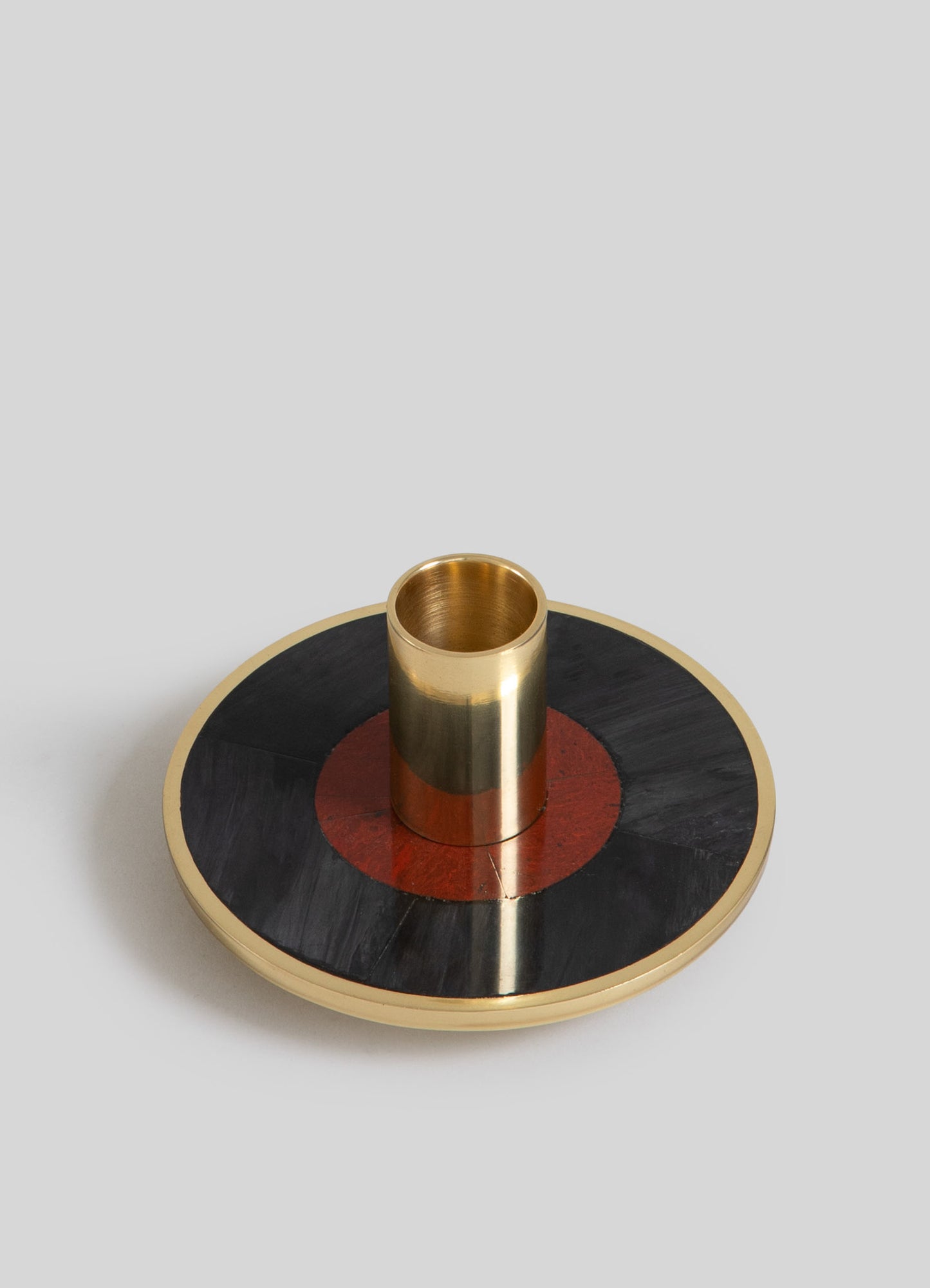 A decorative candlestick holder featuring a round wooden base with a geometric pattern in black and reddish-brown, topped with a cylindrical brass holder in a polished gold finish.