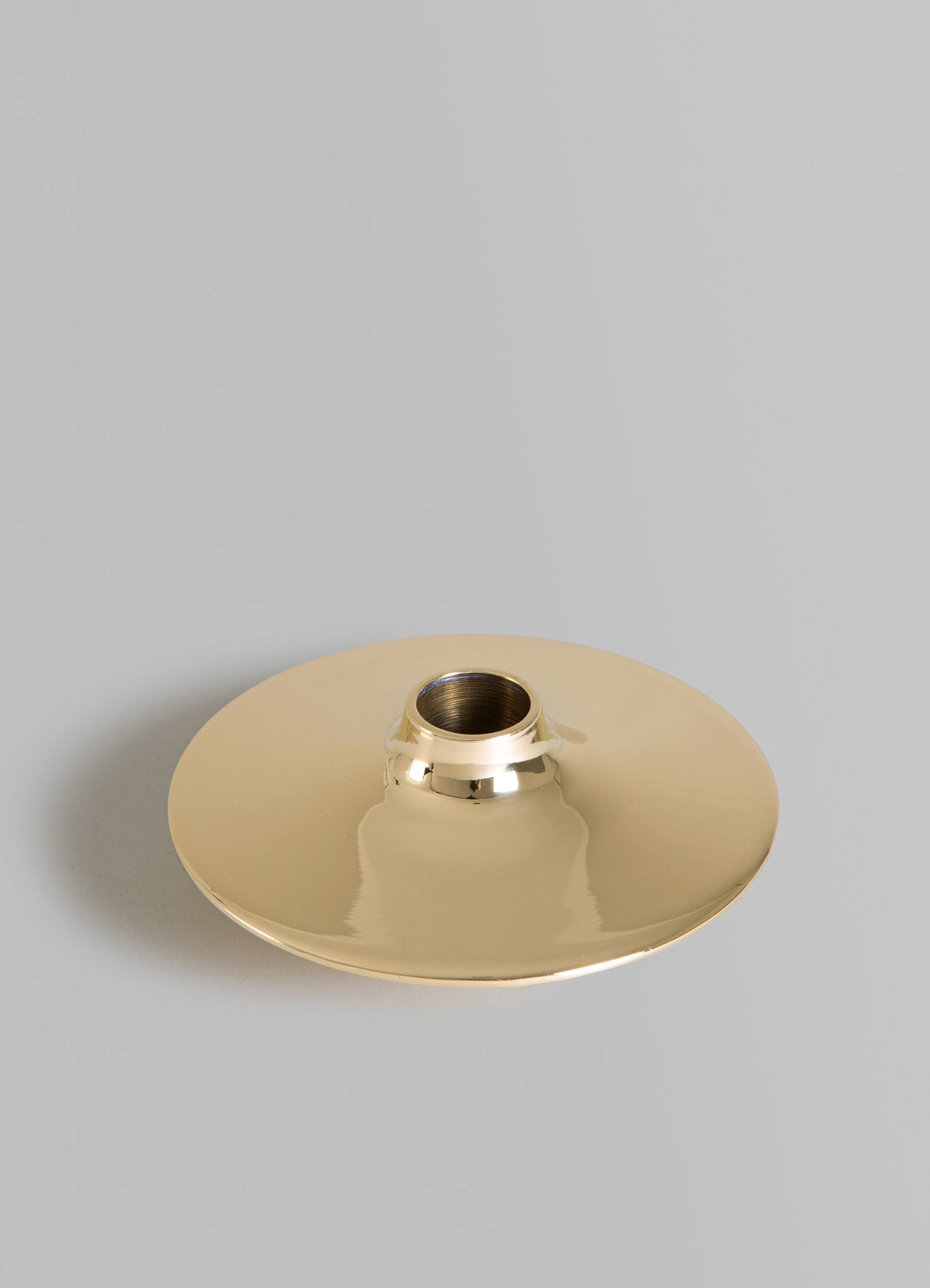 Brass candle holder with a round, flat base and a central cylindrical socket, featuring a polished, reflective surface in metallic gold tones.