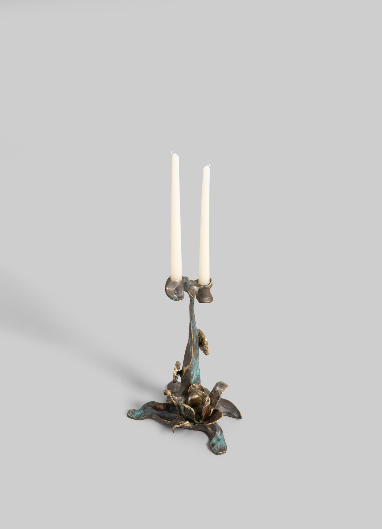 Bronze candelabrum with a sculptural, organic design featuring three candle holders, paired with white taper candles, set against a plain gray background.
