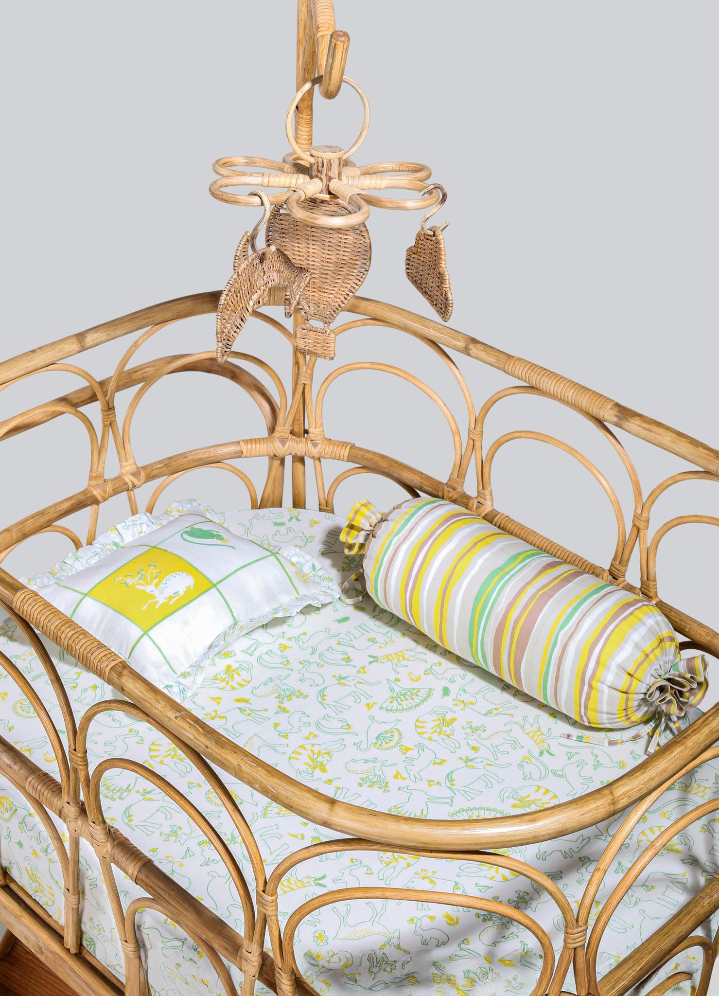 Rattan crib with a curved, open-frame design and a natural wood finish, featuring a canopy with decorative woven leaves and a hanging ring. Bedding includes a white fitted sheet with a colorful dinosaur print, a ruffled pillow with a yellow crest motif, and a cylindrical bolster pillow with pastel-striped fabric.