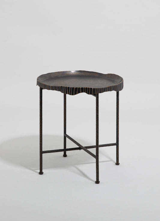 A small side table featuring a black metal frame with four slender, hammered-texture legs and a round, irregularly shaped tabletop with a scalloped edge. The tabletop appears to be made of metal with a dark, matte finish.