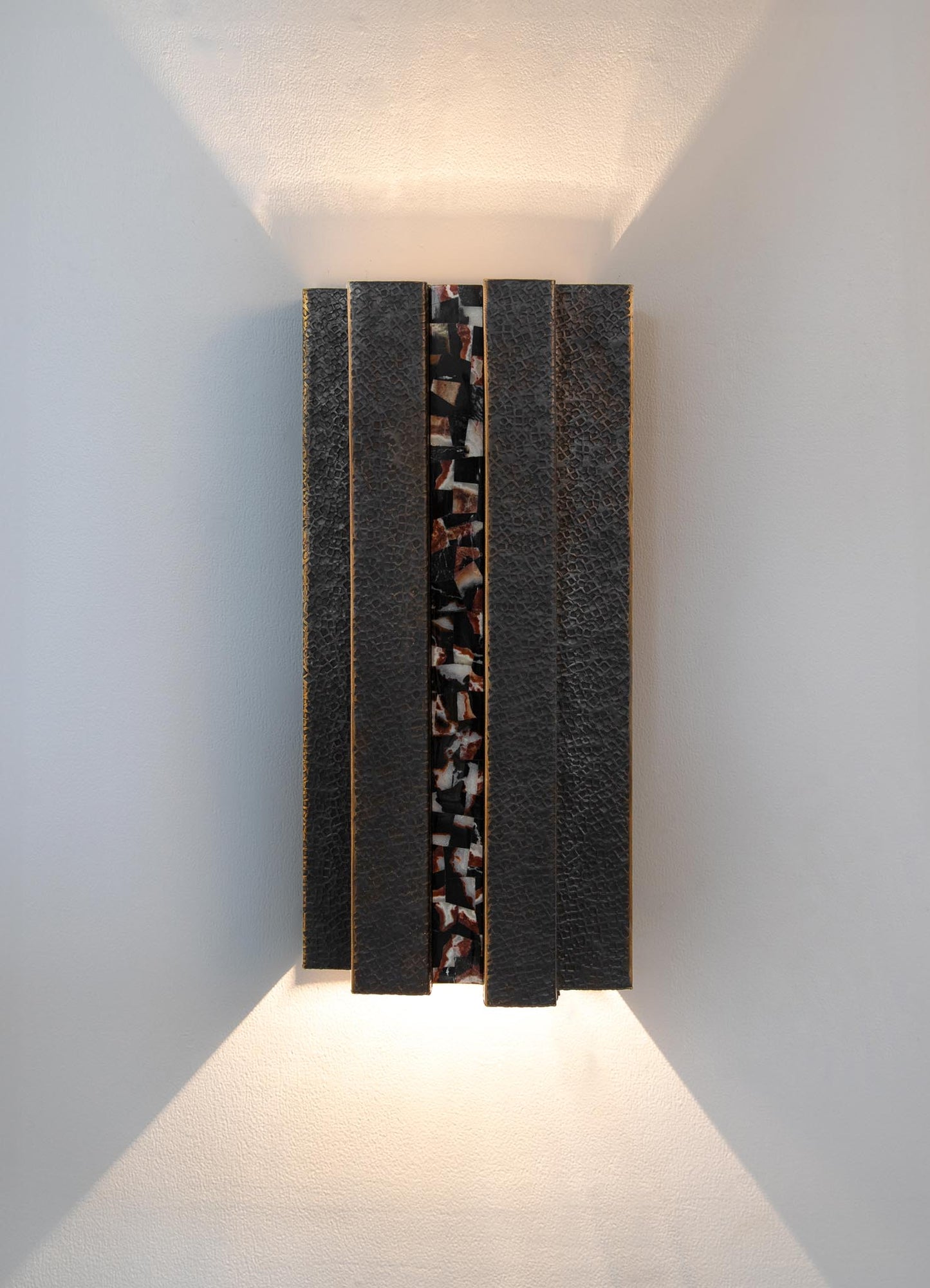 A decorative wall-mounted light fixture with a rectangular shape, featuring a textured, dark-colored surface with embedded mosaic-like patterns in earthy tones, cast in a contemporary style.