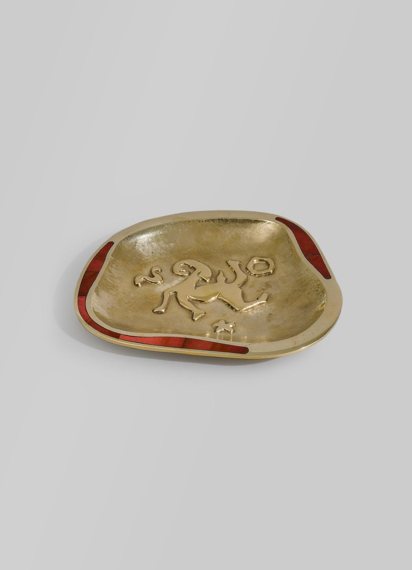 A decorative gold-colored dish with an irregular shape and a raised, textured surface featuring abstract symbols. The dish has red accents along the edges, adding a contrasting detail to the overall design.