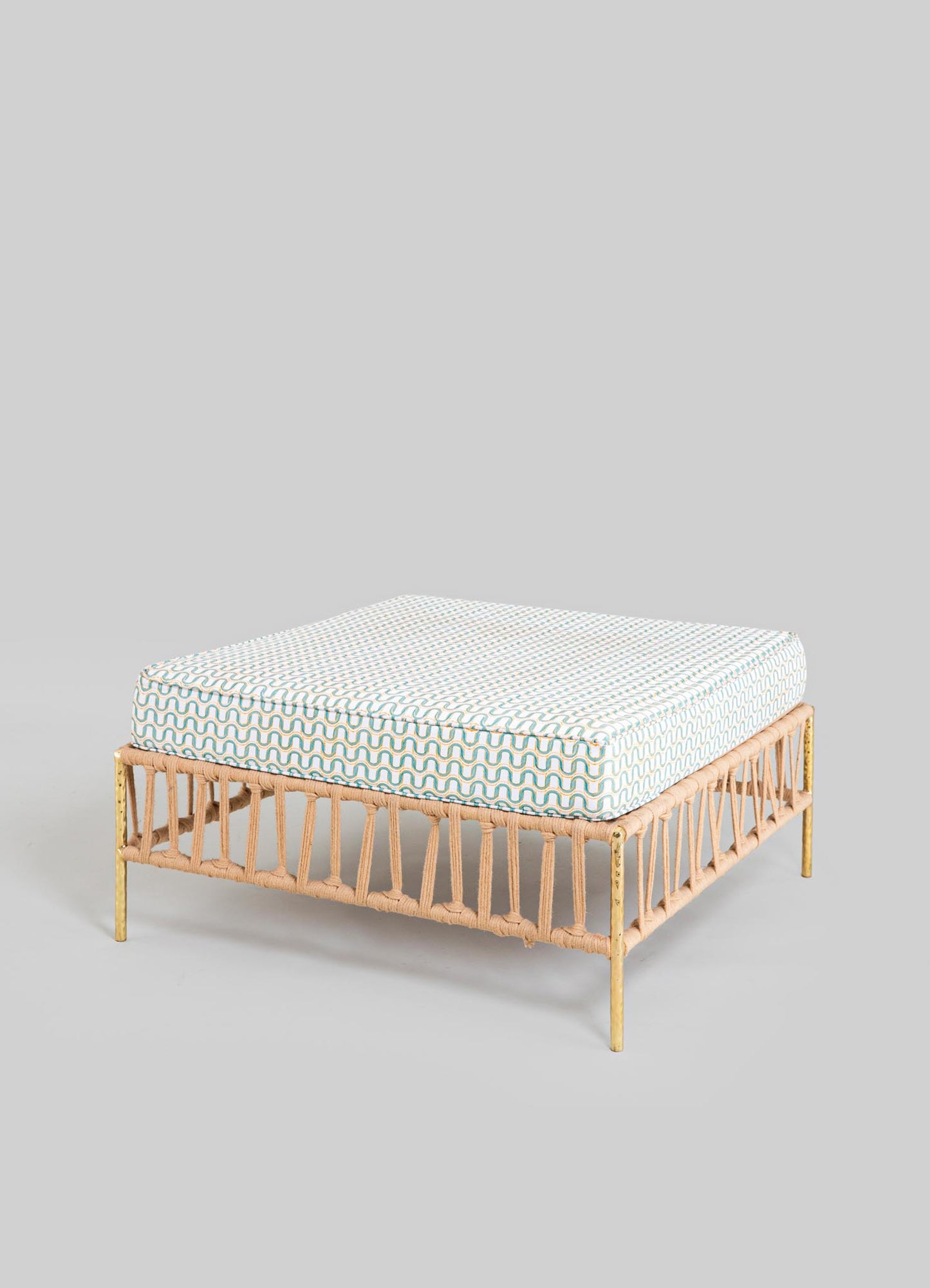 Wicker ottoman with a brass finish on the legs and a cushioned top covered in a patterned fabric featuring wavy blue, beige, and white lines. The design has a contemporary style with an airy, handcrafted look.