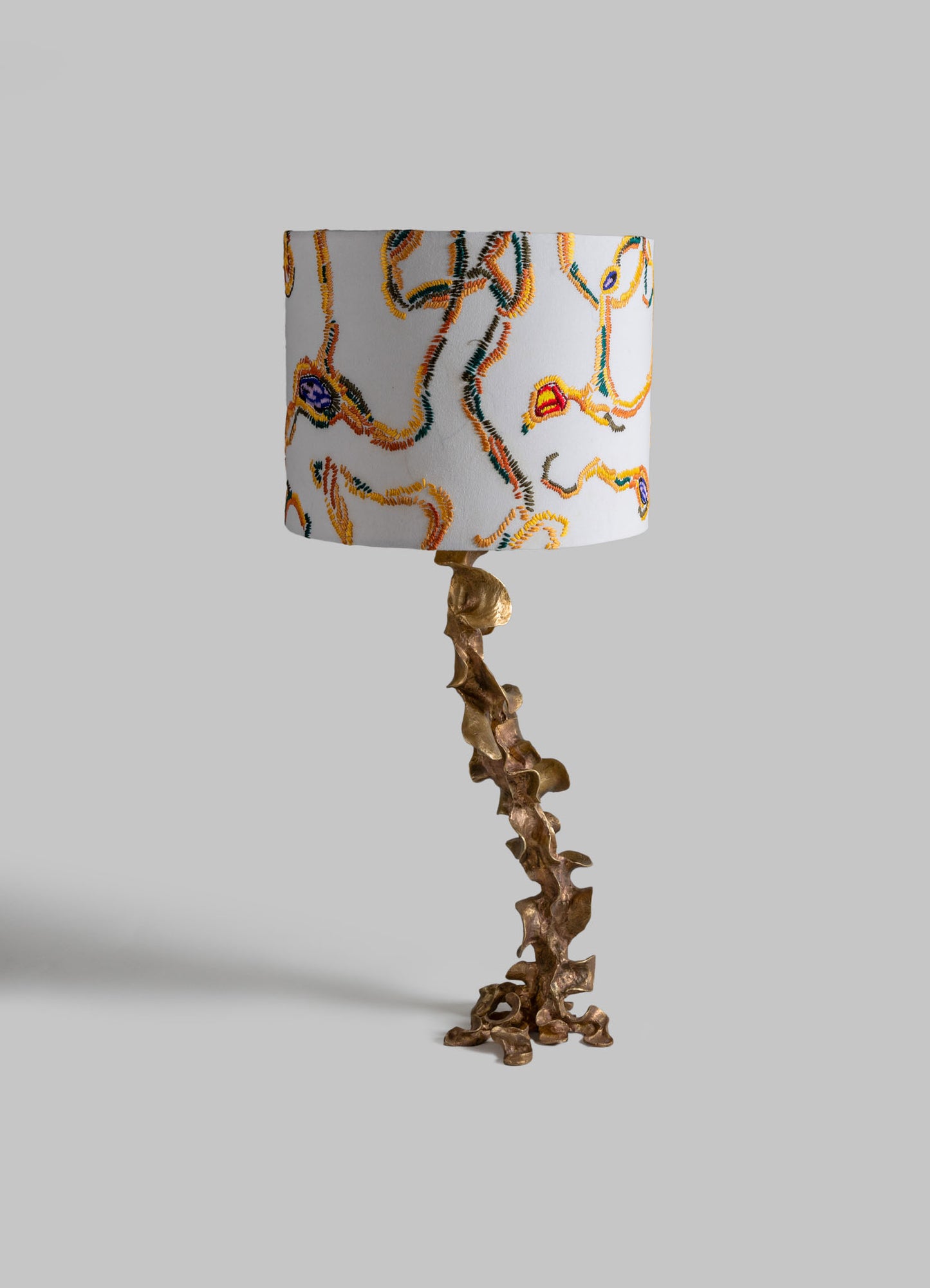 Table lamp with a sculptural, irregular bronze base and a cylindrical fabric lampshade featuring an embroidered abstract, colorful pattern against a plain background.