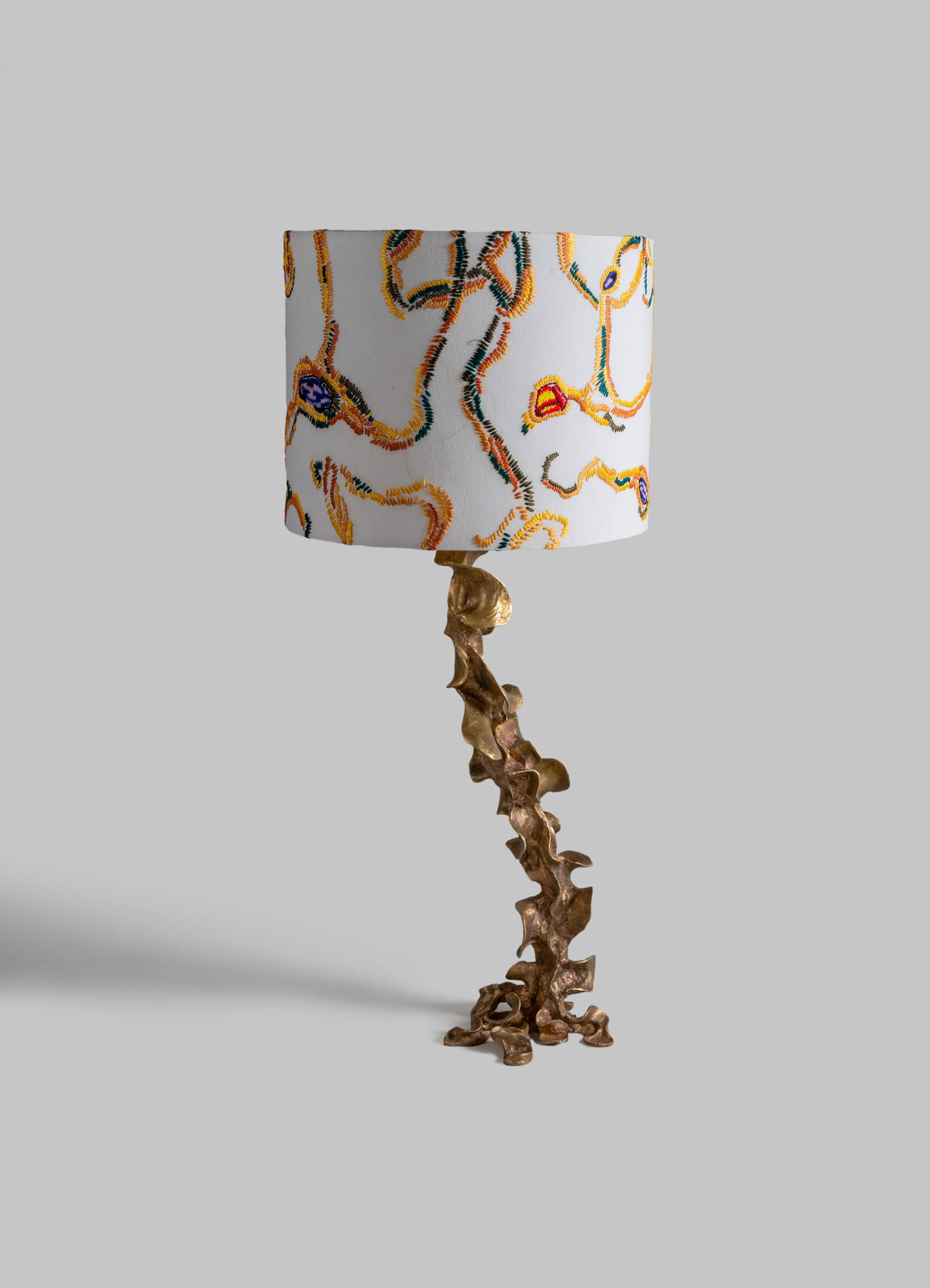 Table lamp with a sculptural, irregular bronze base and a cylindrical fabric lampshade featuring an embroidered abstract, colorful pattern against a plain background.