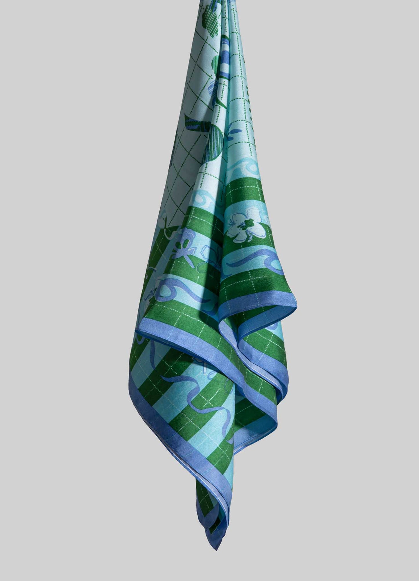 A fabric towel with a floral and geometric pattern in shades of green, blue, and white, edged with a solid blue border. The towel is draped in a vertical position against a plain background.