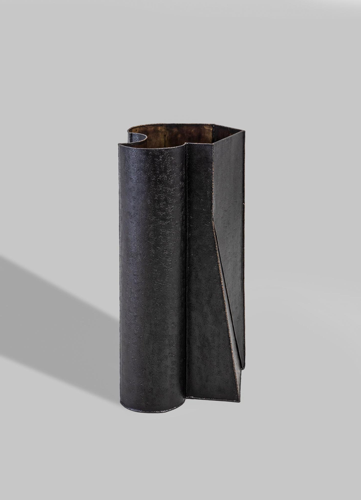 A tall, cylindrical metal vase with a matte black finish, featuring an asymmetrical, folded design element on one side. The texture appears slightly rough, and the interior reveals a rusted or aged metallic surface.