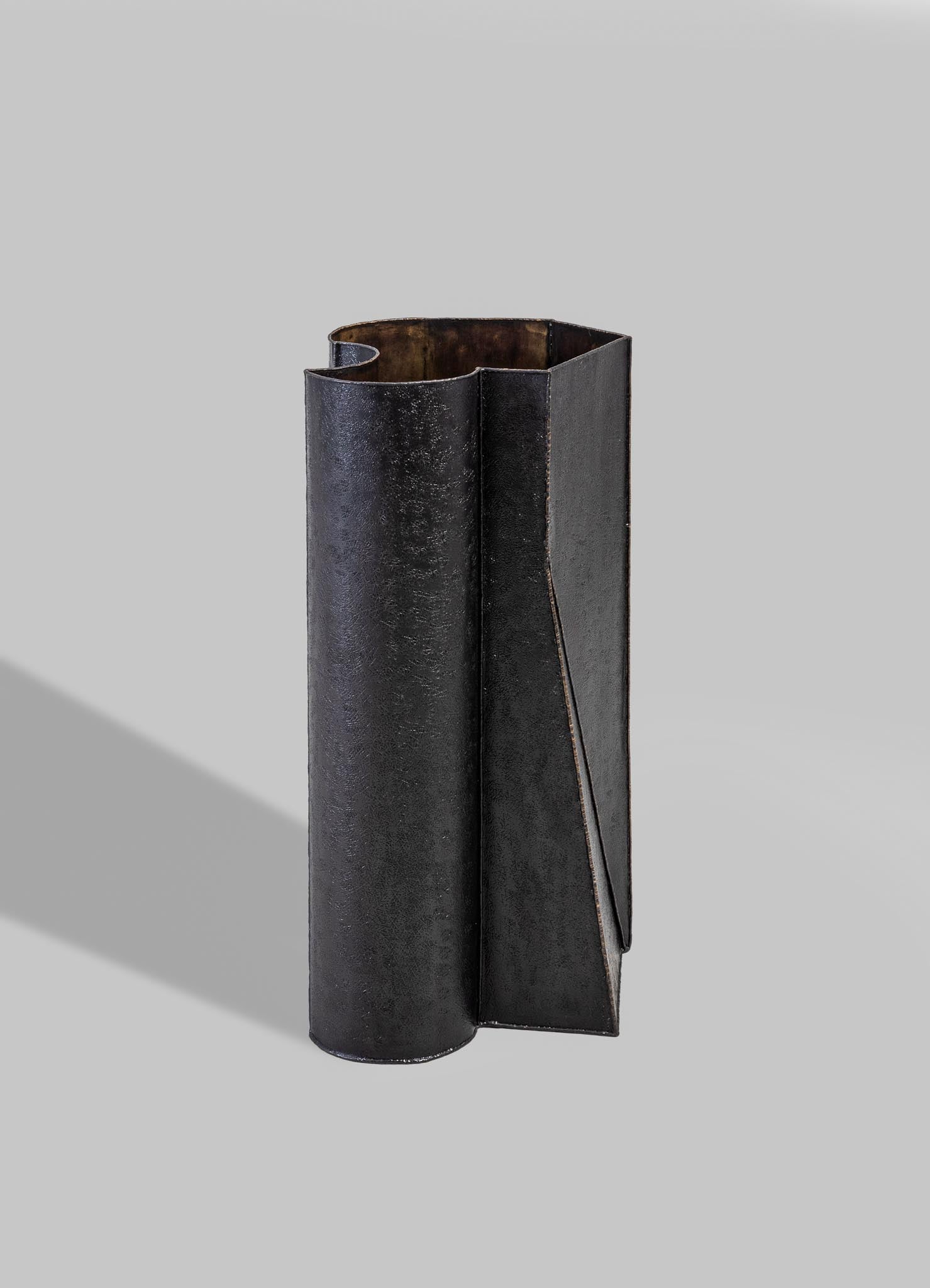 A tall, cylindrical metal vase with a matte black finish, featuring an asymmetrical, folded design element on one side. The texture appears slightly rough, and the interior reveals a rusted or aged metallic surface.