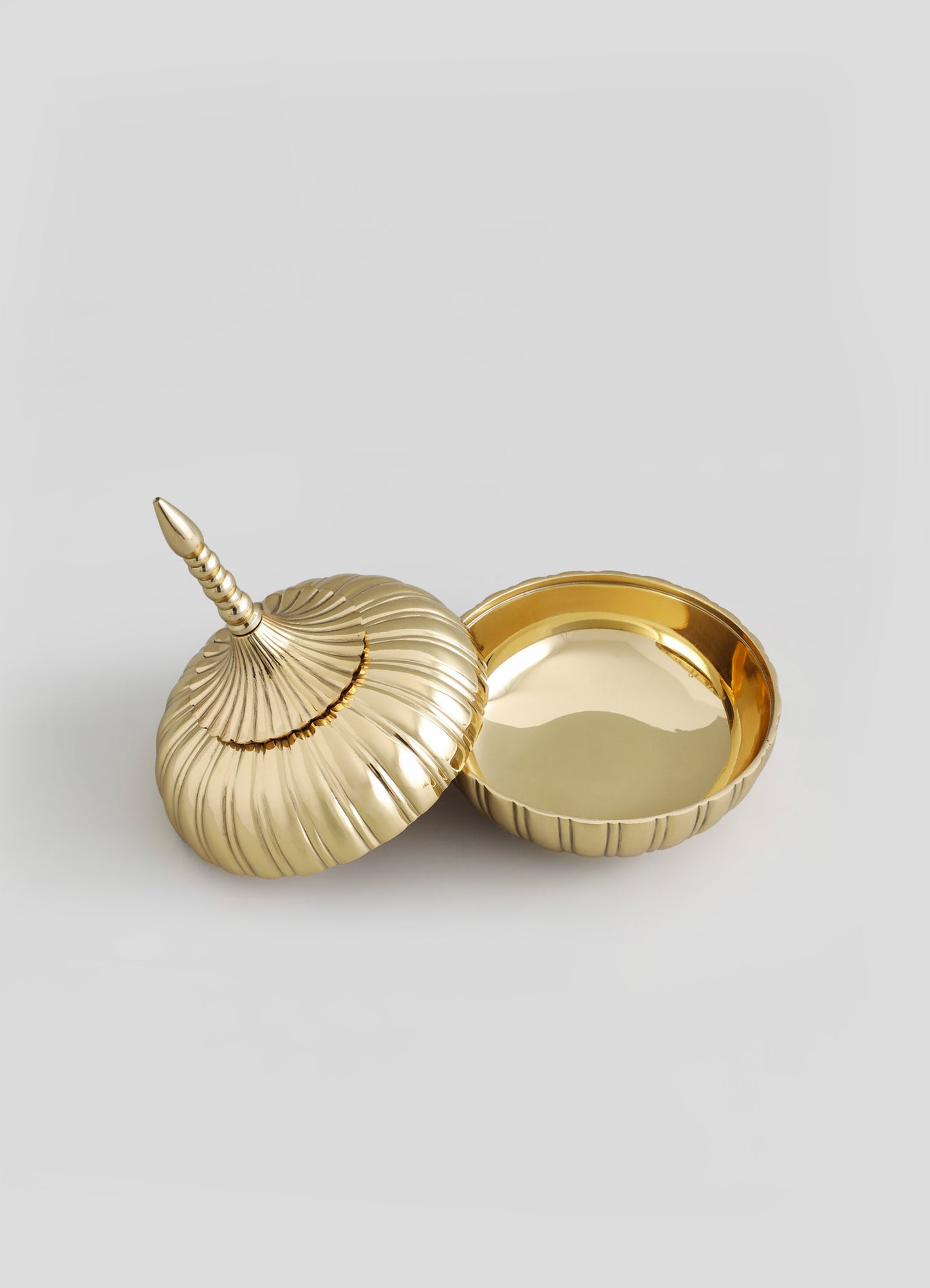 A gold-toned decorative tray and lid resembling a seashell with a pointed, beaded finial, made of metal with a reflective, polished surface. The design features embossed ridges and a smooth interior, combining coastal motifs with modern luxury style.