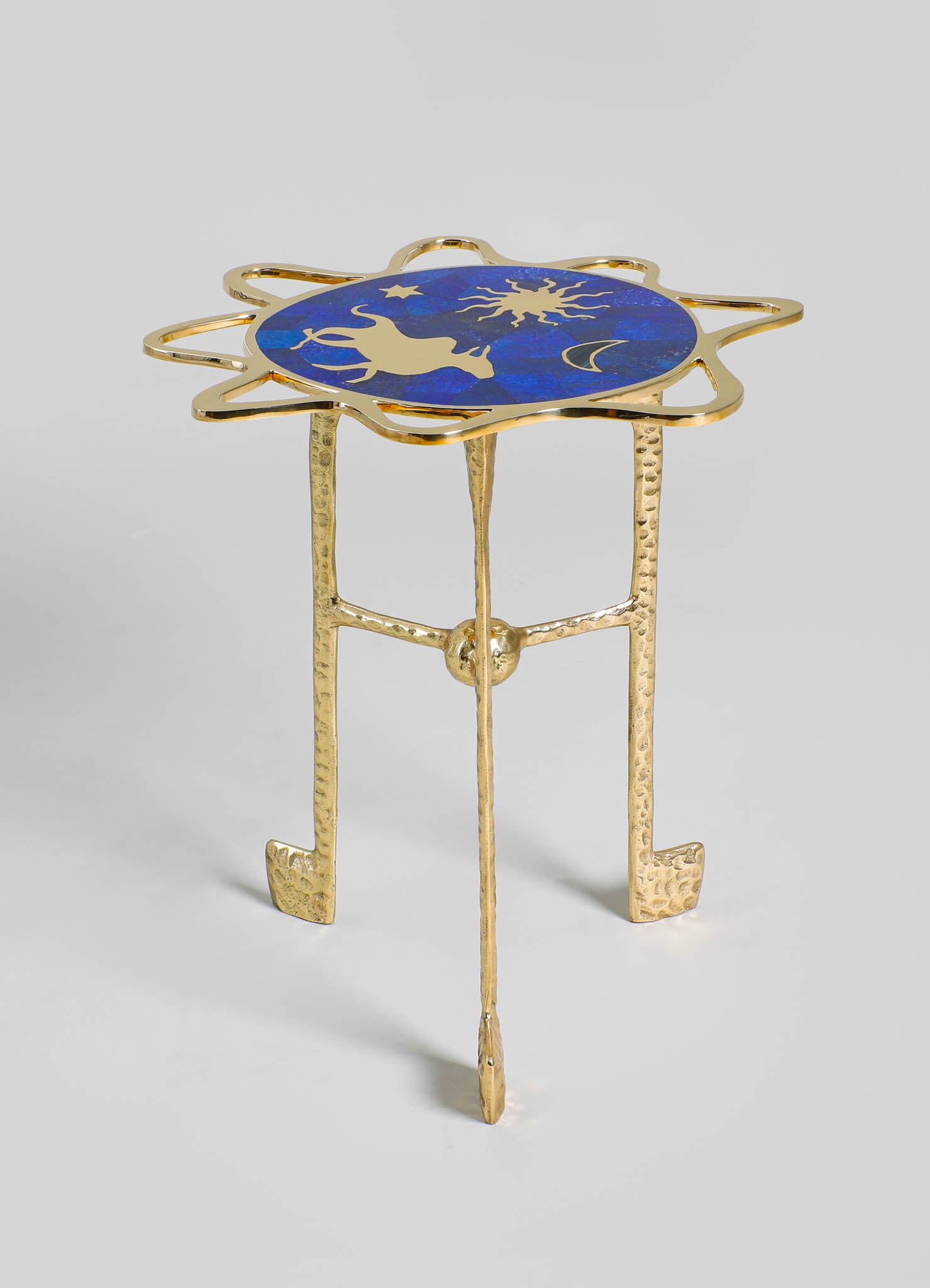A decorative side table with a gold-finished metal frame and textured legs, featuring a round top with a celestial motif in blue, gold, and black. The top displays abstract sun, moon, and star designs on a dark blue background.