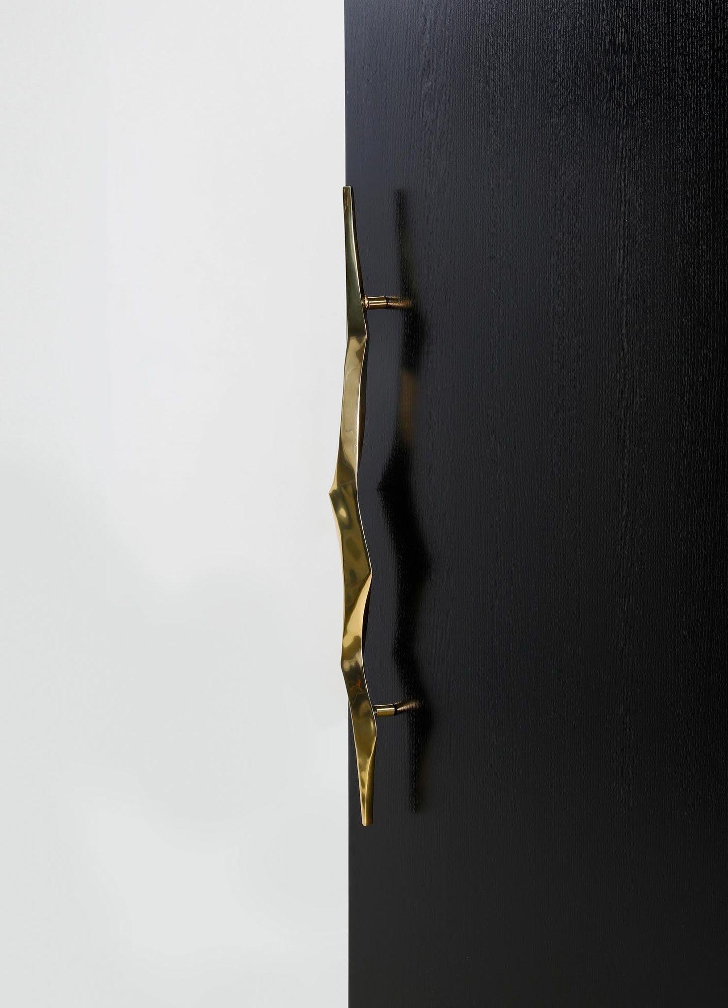 A metallic gold door handle with a wavy, sculptural design mounted on a black textured door, contrasting with a plain white wall in the background.