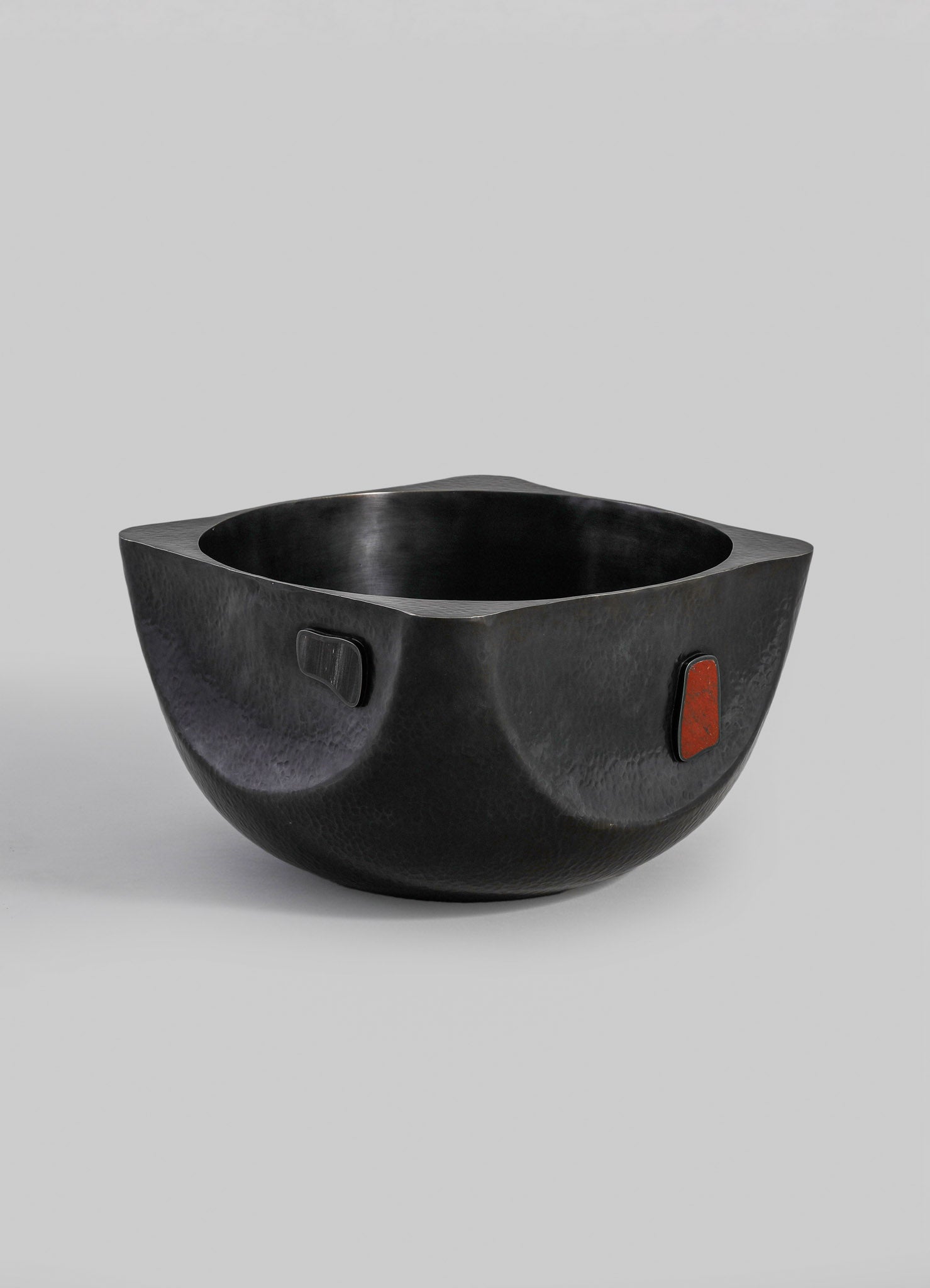 A black hammered metal bowl with a rounded, organic shape and small rectangular accents in red and black on the sides.