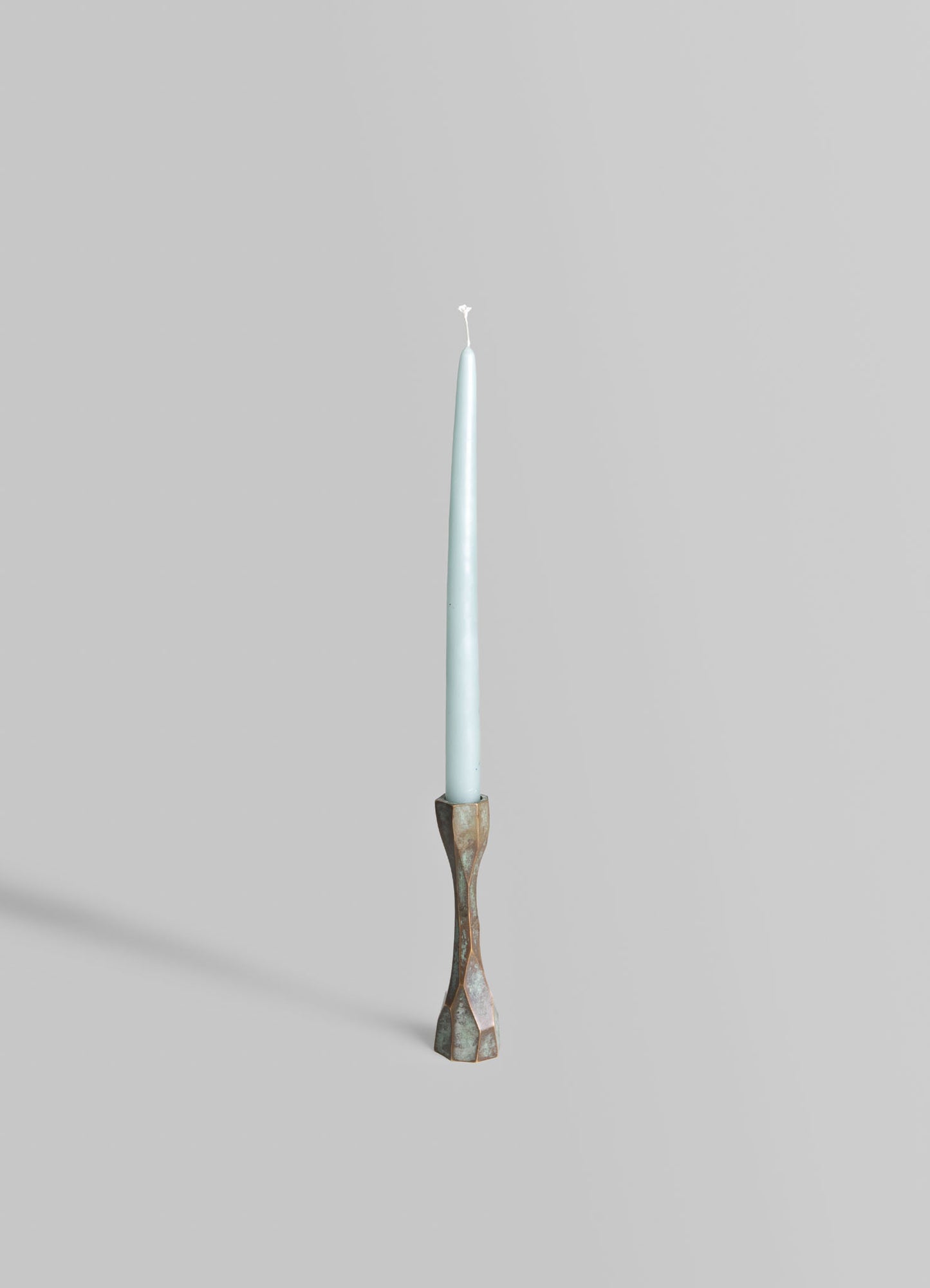 A tall, slender candle with a light blue color and a white wick, supported by a textured, metallic bronze candle holder with an irregular, artistic shape.
