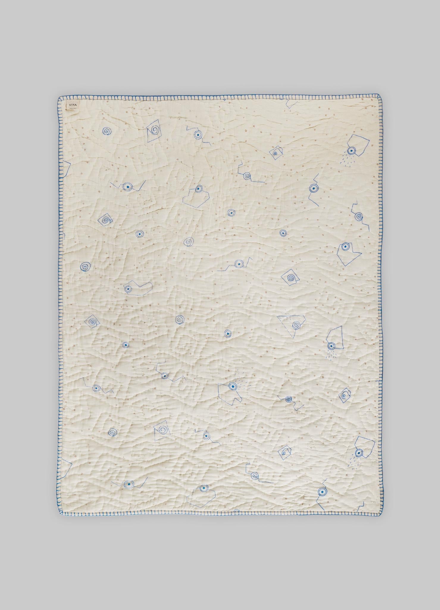 A quilt made of cream-colored fabric with a textured, slightly crinkled surface featuring blue embroidered geometric motifs and a border of evenly spaced blue stitches.