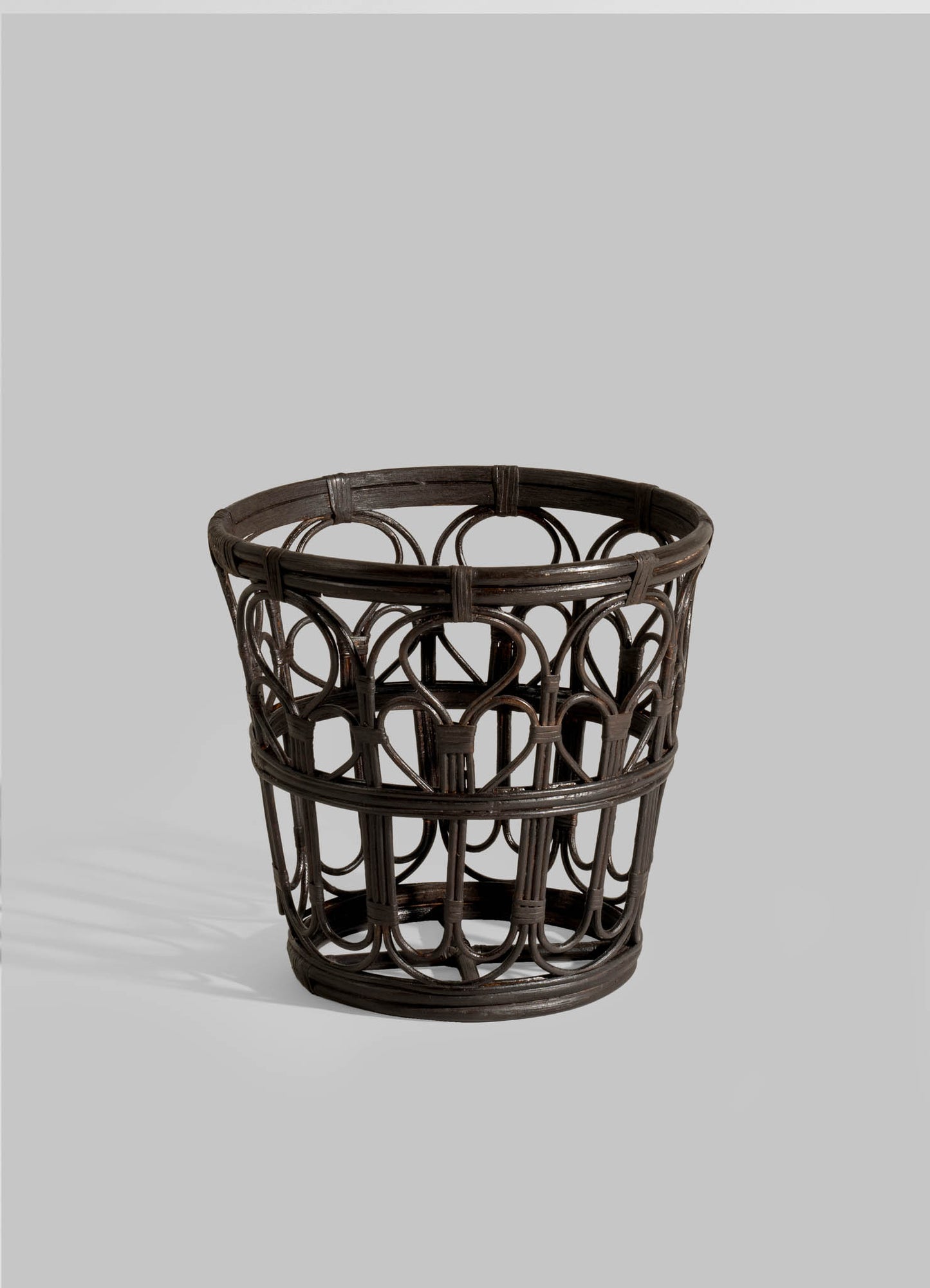 The VIYA Manipur Champagne Bucket is a dark brown, round wicker piece with an open geometric pattern of curved and straight rattan, perfect for home decor. Displayed against a plain light gray background.
