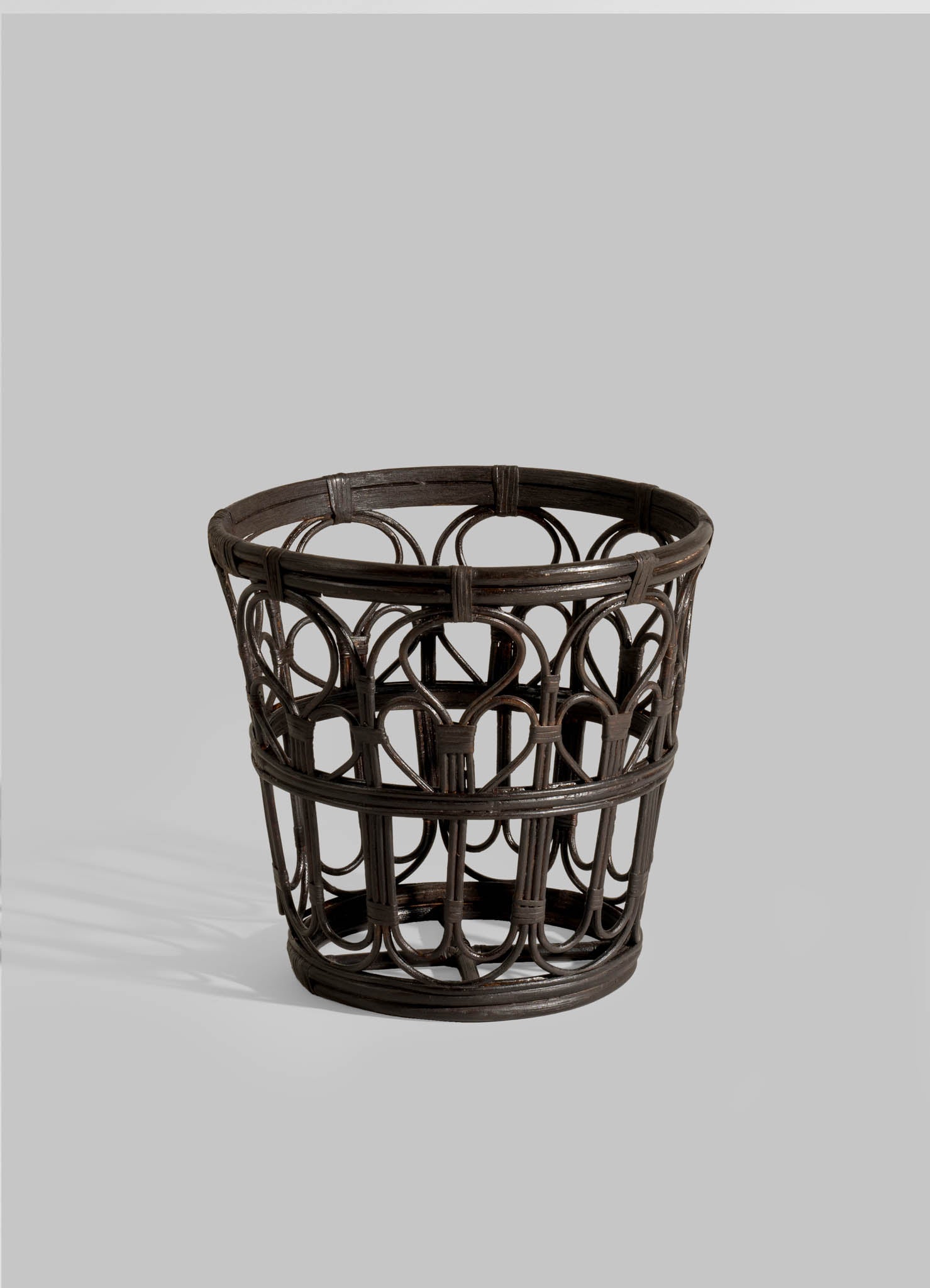The VIYA Manipur Champagne Bucket is a dark brown, round wicker piece with an open geometric pattern of curved and straight rattan, perfect for home decor. Displayed against a plain light gray background.
