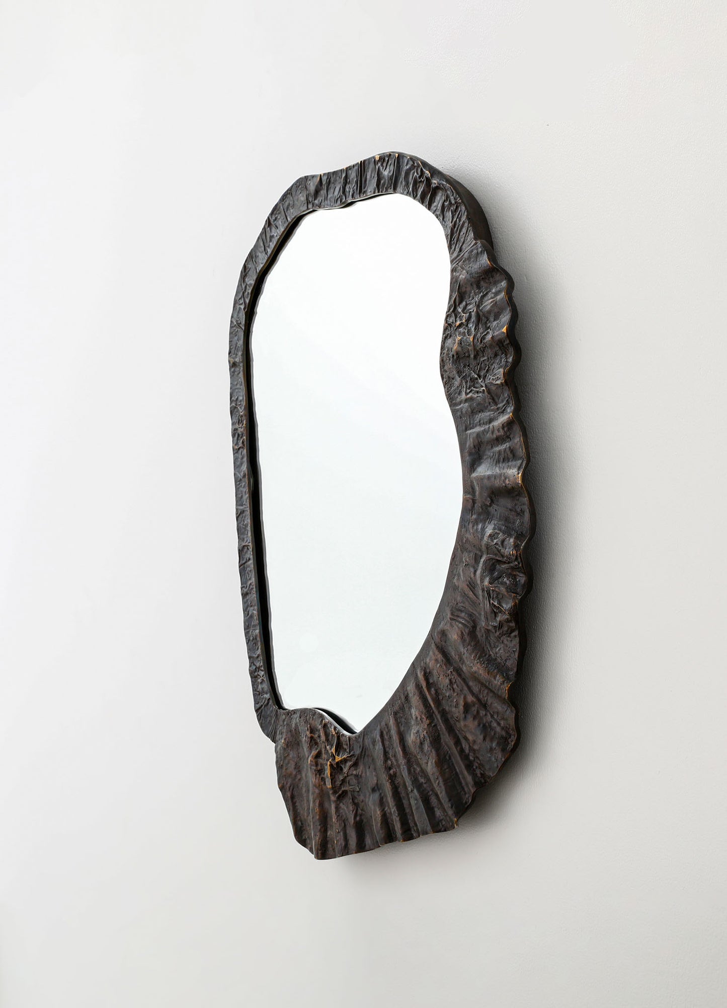 A mirror with a textured, irregularly shaped wooden frame featuring a dark finish, mounted on a plain white wall. The frame has a wavy, organic silhouette that emphasizes natural design elements.