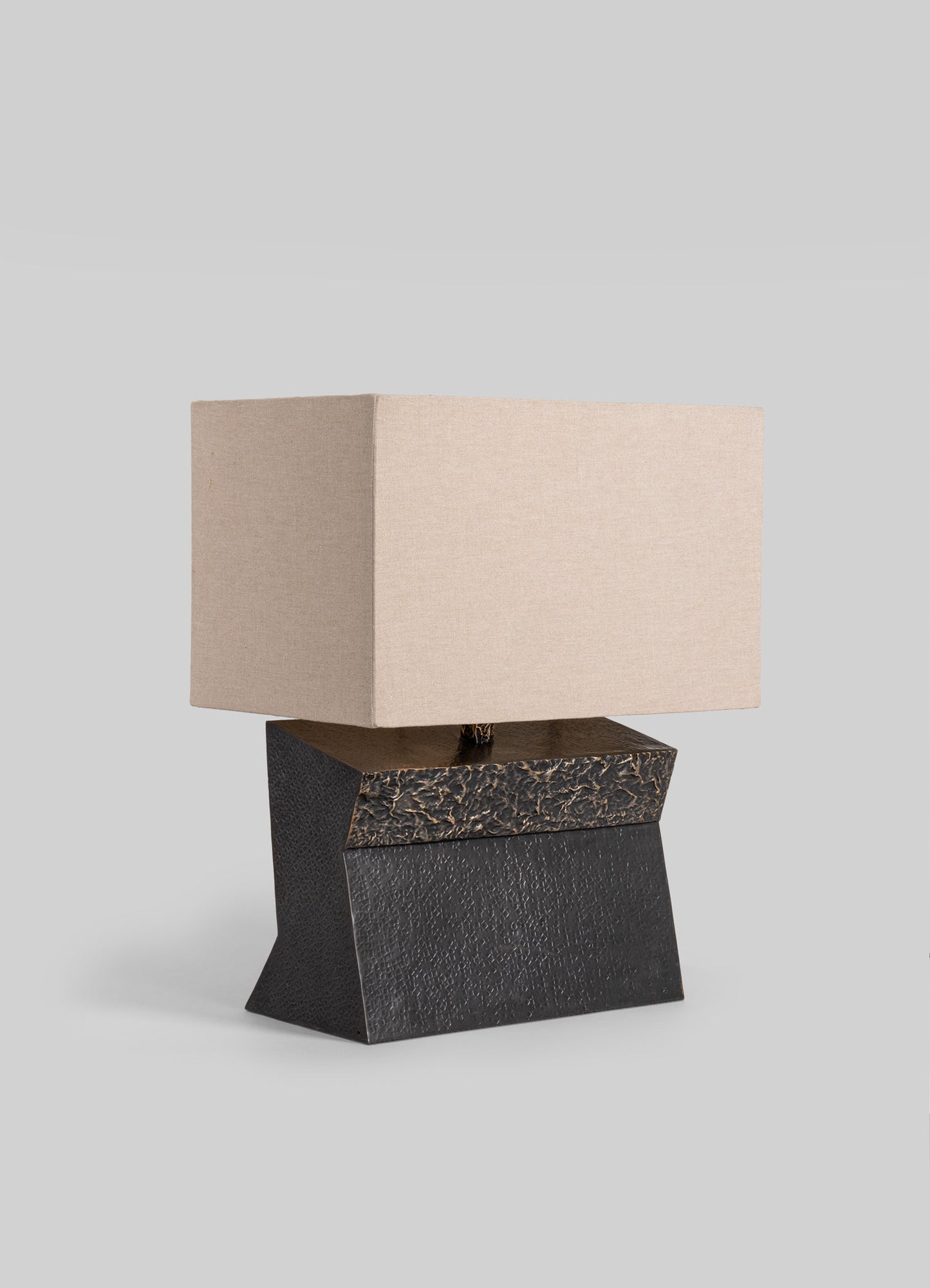 A table lamp featuring a textured black base with geometric shapes and a beige fabric shade, set against a plain light gray background.