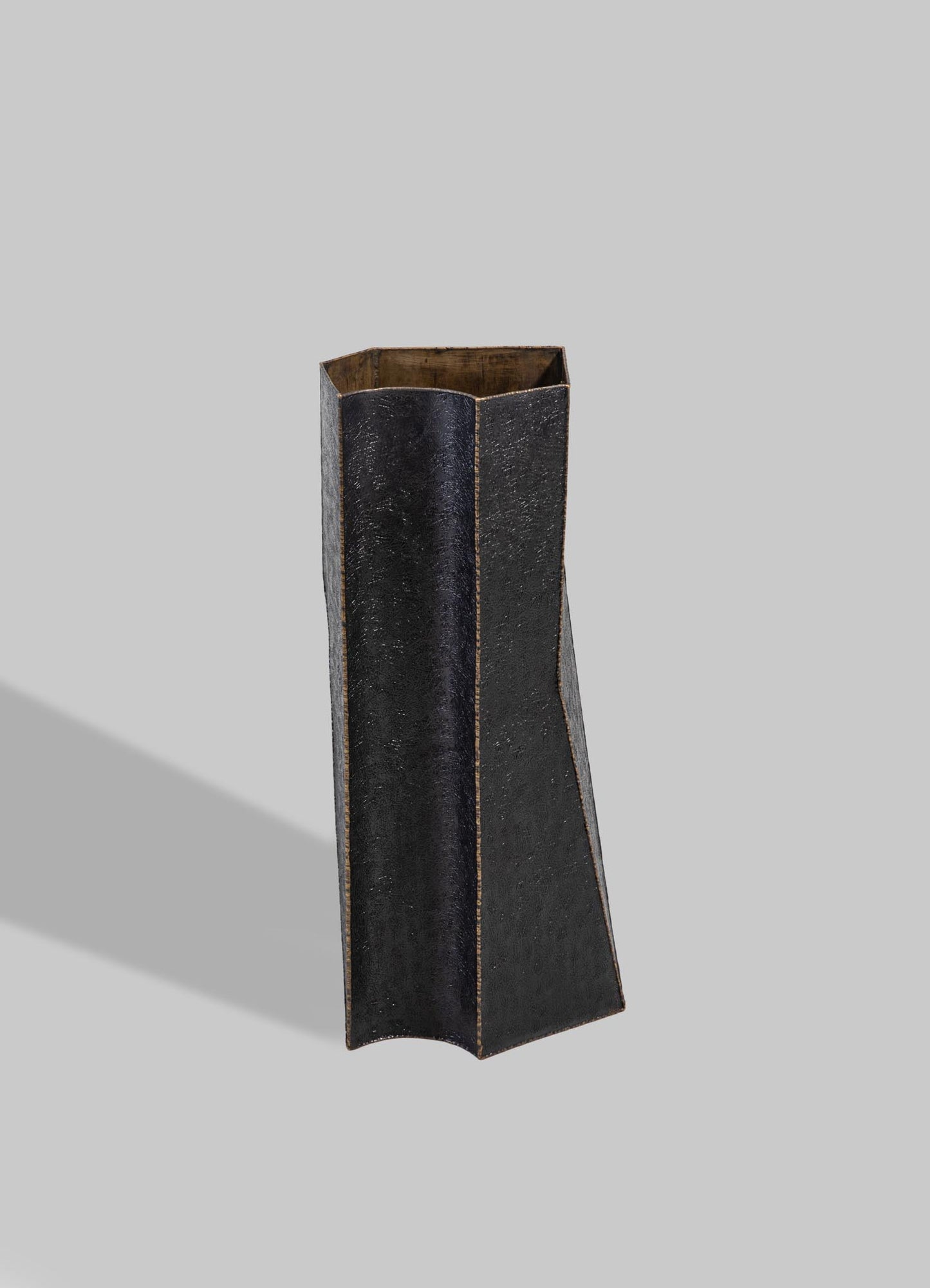 A tall, sculptural vase made of dark, textured material with a matte black finish and a narrow, slightly flared opening. The interior reveals natural wood tones, contrasting with the exterior's modern aesthetic.