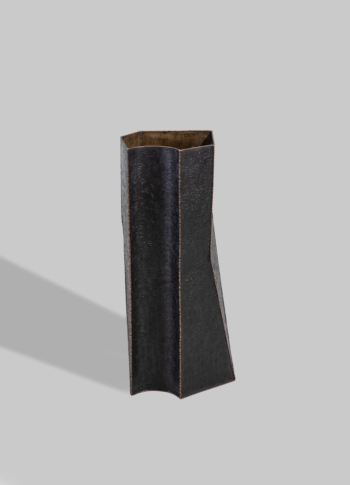 A tall, sculptural vase made of dark, textured material with a matte black finish and a narrow, slightly flared opening. The interior reveals natural wood tones, contrasting with the exterior's modern aesthetic.