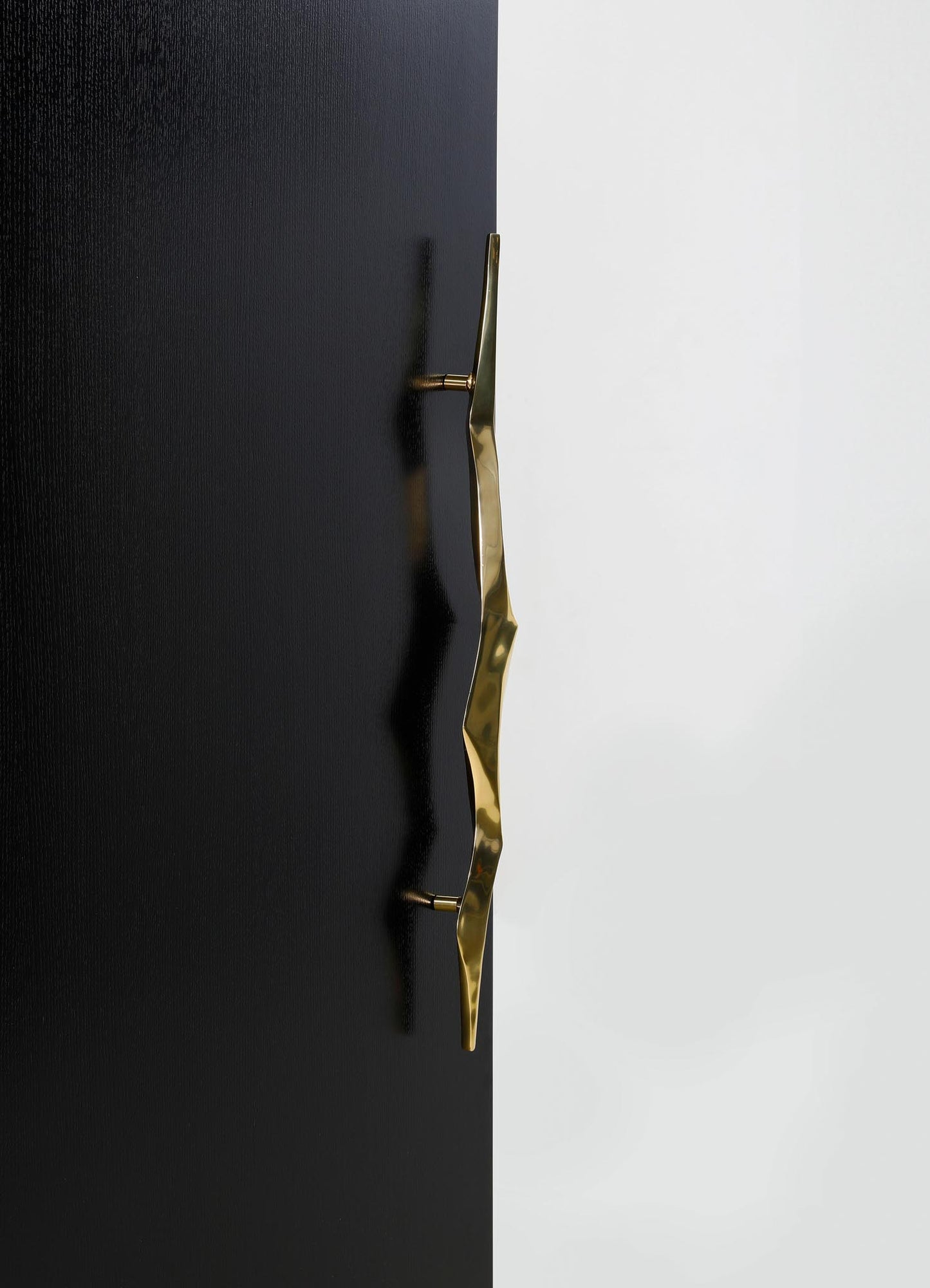 A black wooden door with a wavy, irregular brass handle mounted in the center, featuring a polished, reflective surface. The handle's design is modern and sculptural, contrasting with the textured finish of the door.