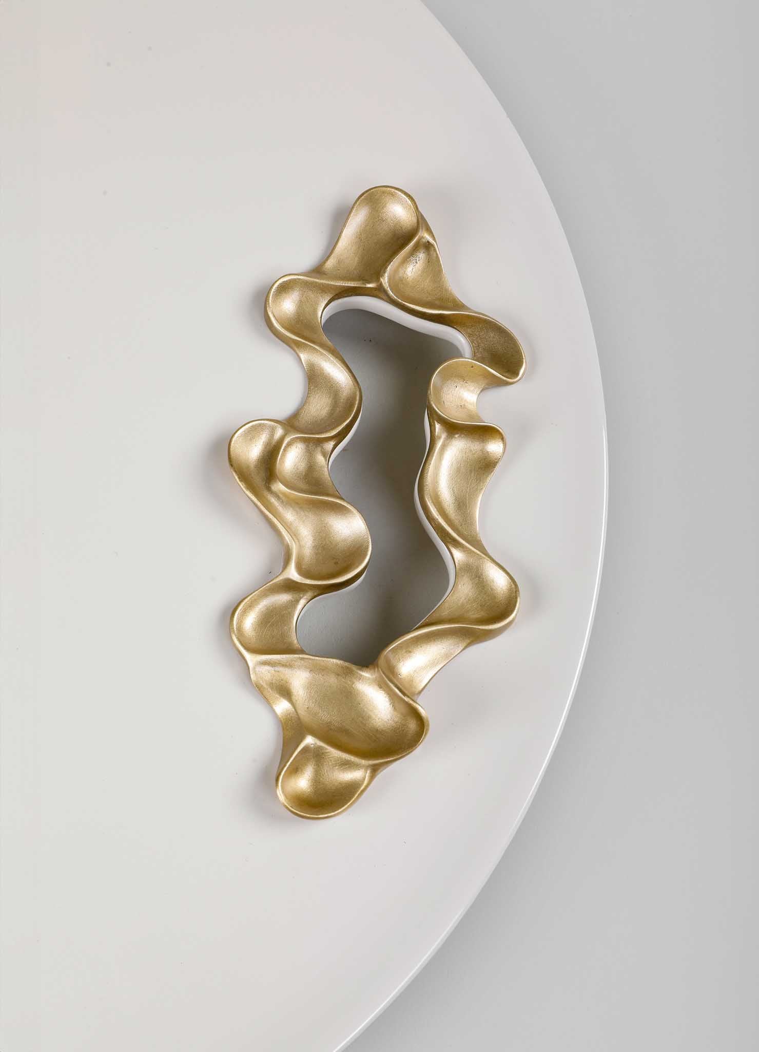 A decorative metal sculpture with an organic, flowing form and a metallic gold finish, displayed on a white surface with a curved edge.