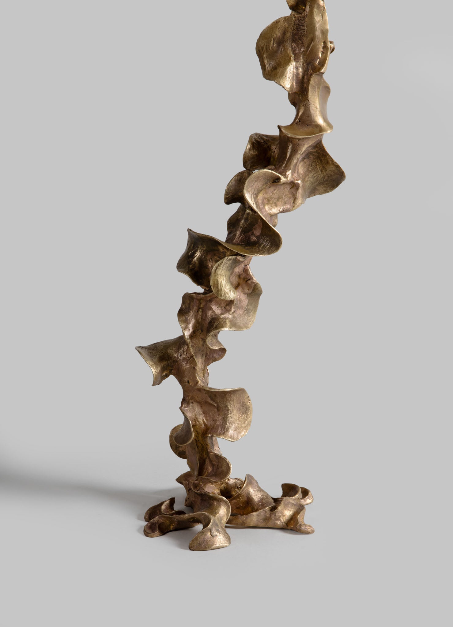 A decorative sculpture made of metal with a polished, reflective surface in a warm bronze tone, featuring an abstract, organic form with flowing, irregular shapes. The sculpture has a contemporary style with a smooth finish and fluid design.
