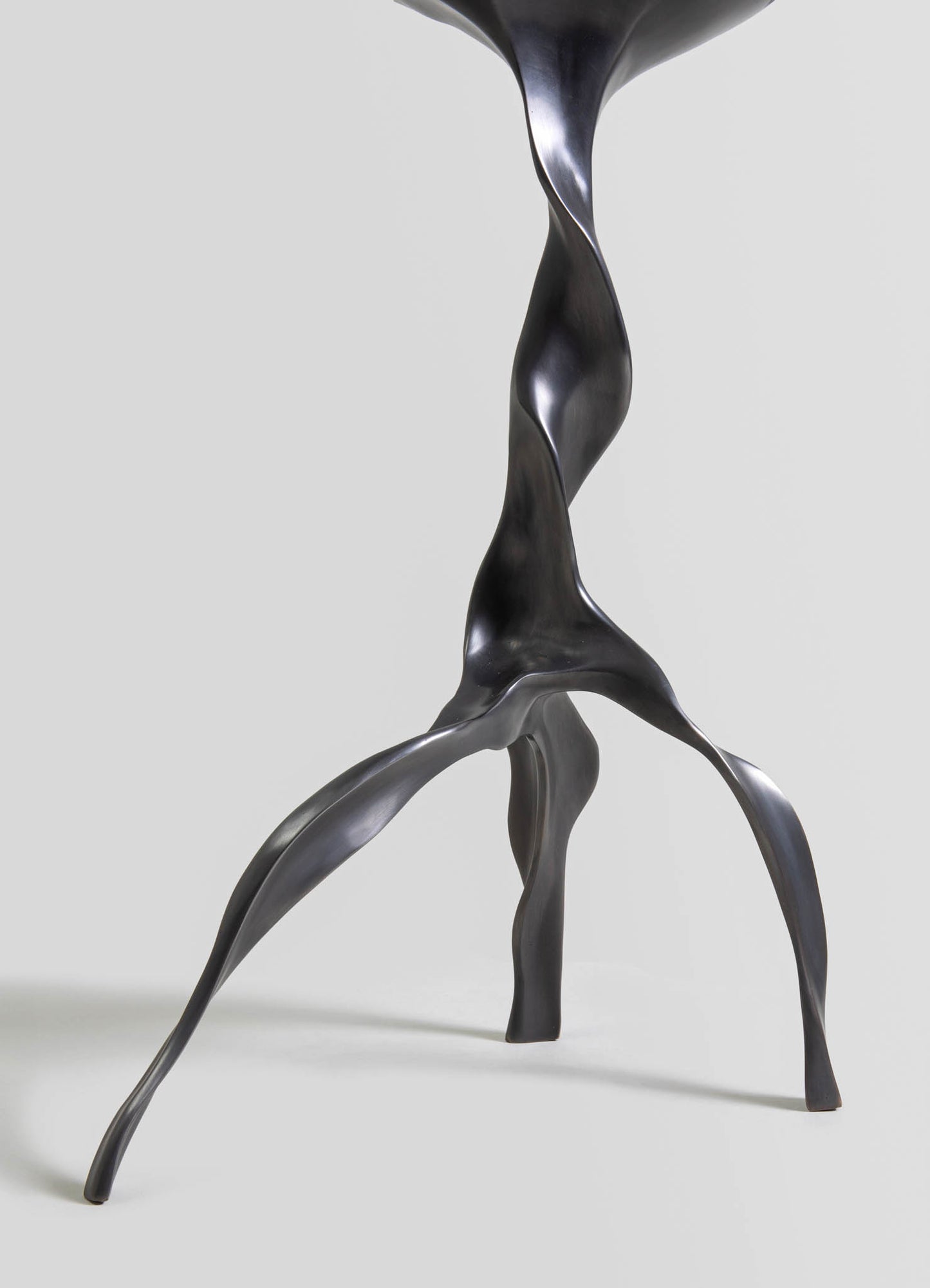 Sculptural table leg made of dark metal with a twisted, organic shape on three angled legs, conveying a contemporary and artistic style.