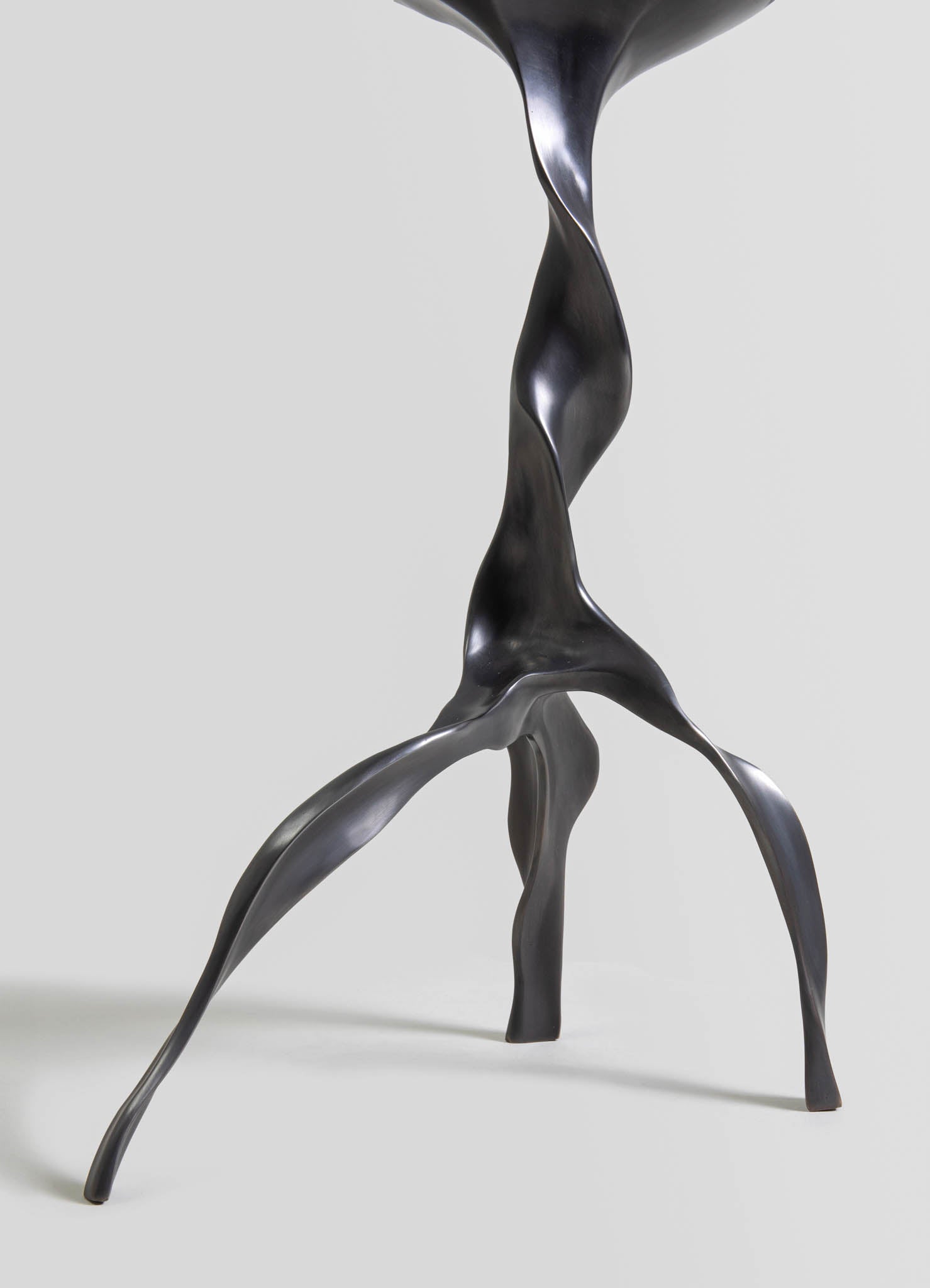 Sculptural table leg made of dark metal with a twisted, organic shape on three angled legs, conveying a contemporary and artistic style.