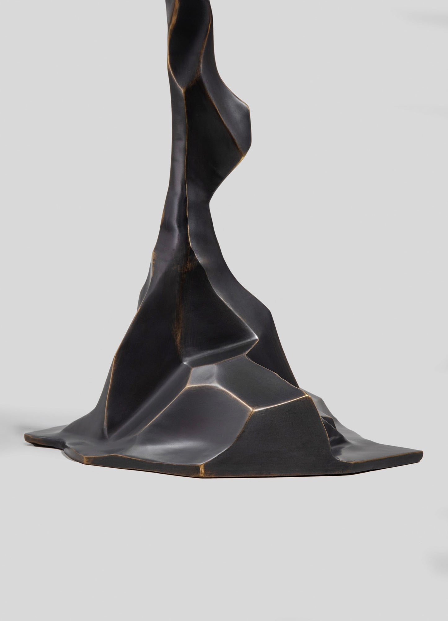 A sculpture made of dark metal with an abstract, flowing form featuring smooth, curved surfaces and sharp edges set against a plain gray background.