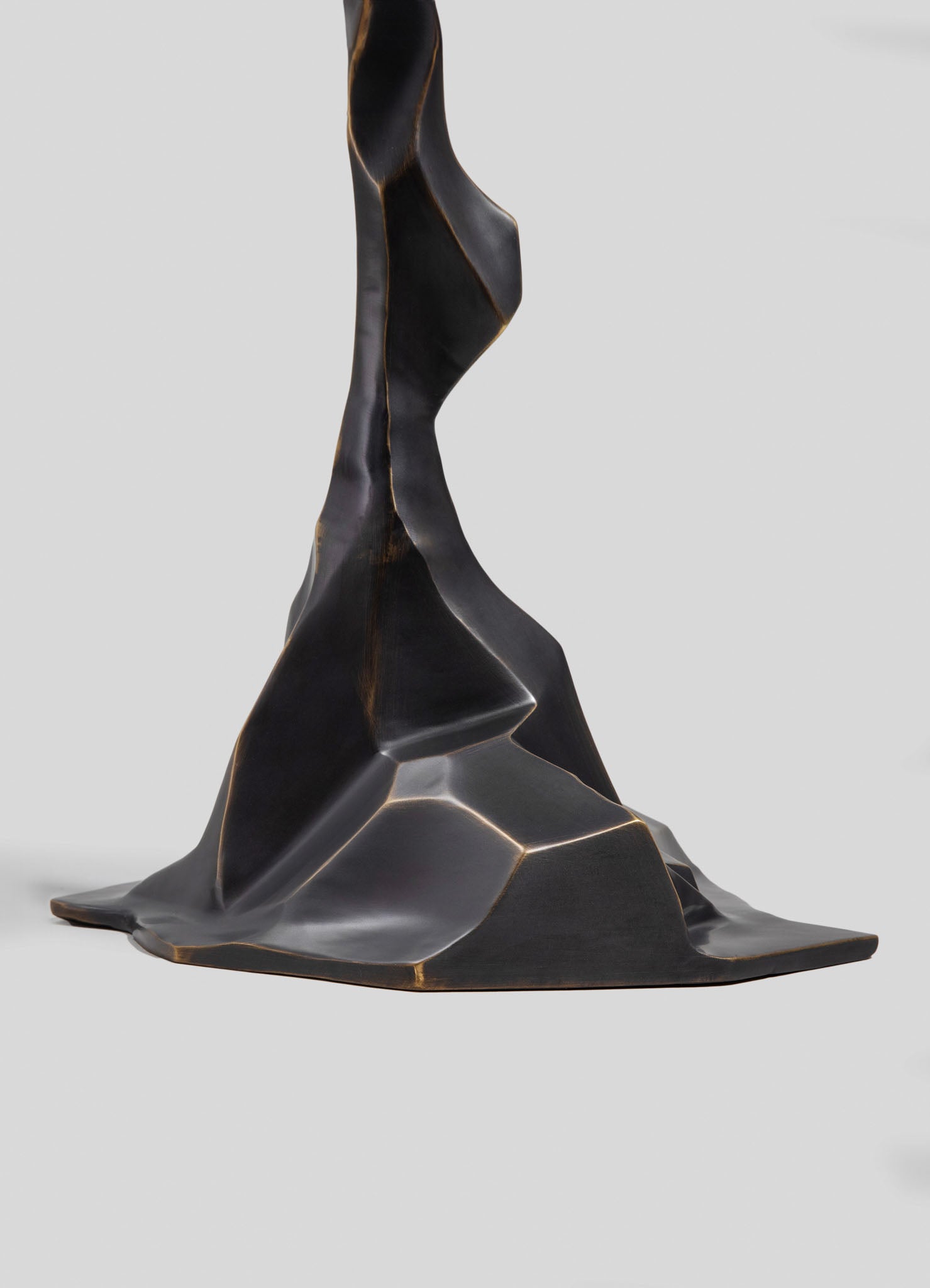 A sculpture made of dark metal with an abstract, flowing form featuring smooth, curved surfaces and sharp edges set against a plain gray background.