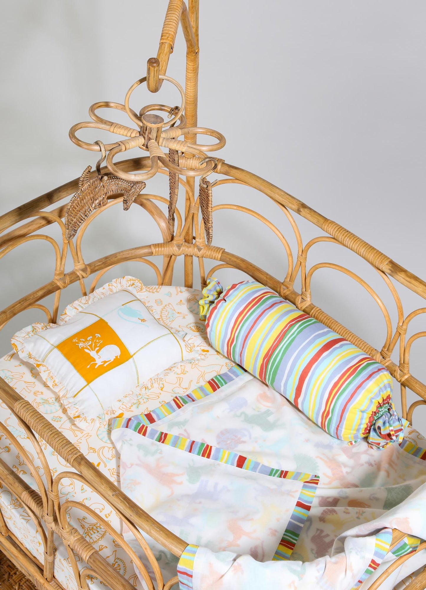 Rattan baby crib with curved and woven design elements, featuring a multicolored striped fabric pillow and white bedding with orange decorative motifs, set against a plain background.