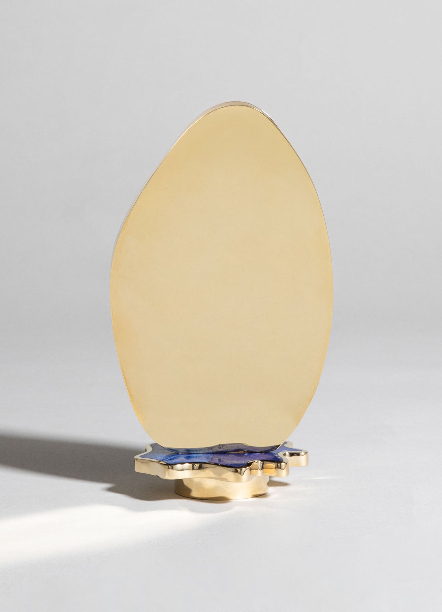A lamp with an oval-shaped, opaque yellow glass shade mounted on a decorative base with metallic and blue accents, set against a plain background. The lamp combines contemporary style with a smooth, minimalist design.