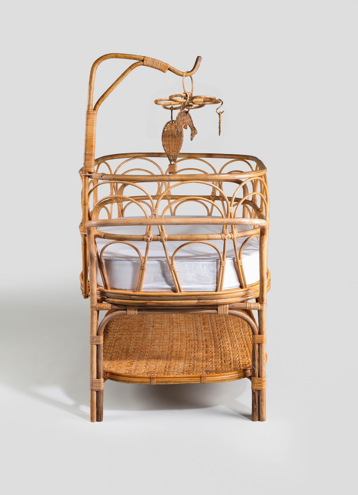 Wicker baby bassinet with a rounded frame, featuring a woven rattan construction and a natural wood finish. The bassinet has a two-tiered design with a padded fabric mattress inside and an attached overhead mobile with rattan decorative elements.