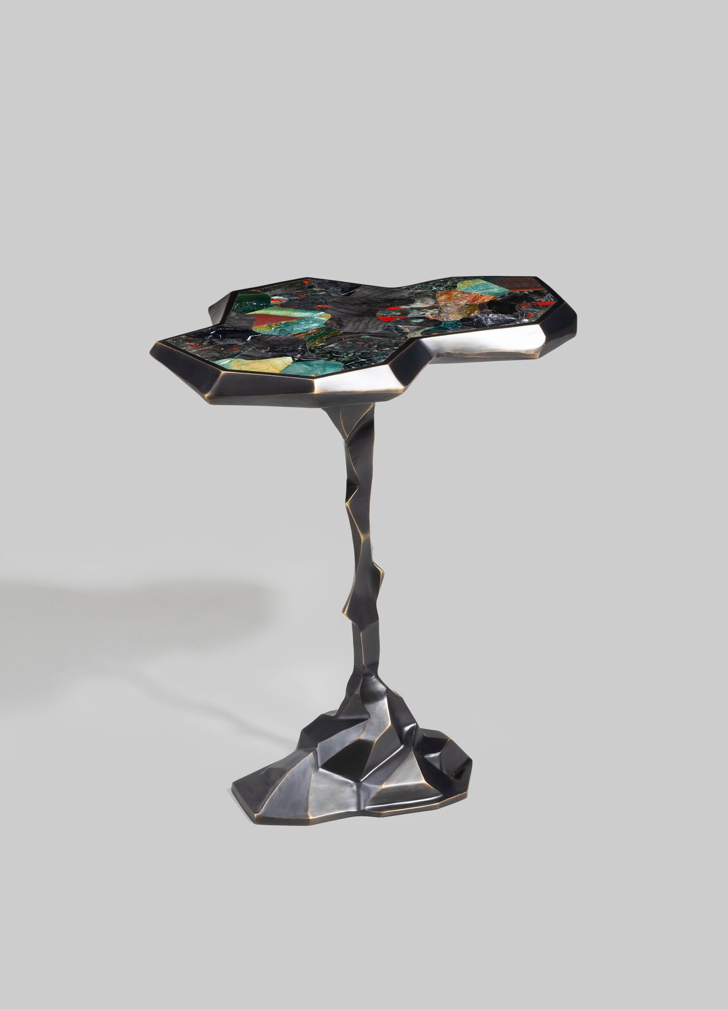 A sculptural side table with a black metal frame featuring a textured, irregular base and a geometric-shaped top inlaid with multicolored stone or glass fragments, creating a mosaic pattern.
