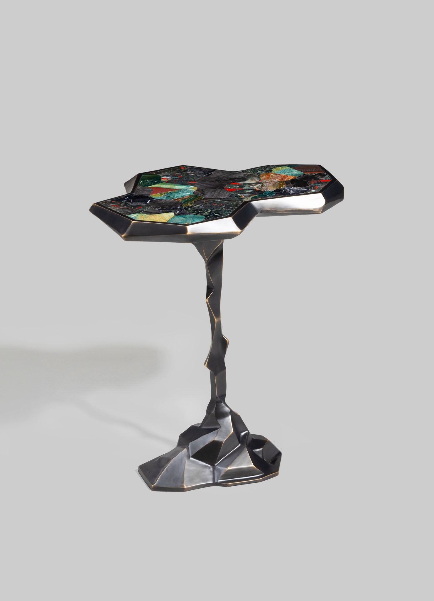 A sculptural side table with a black metal frame featuring a textured, irregular base and a geometric-shaped top inlaid with multicolored stone or glass fragments, creating a mosaic pattern.
