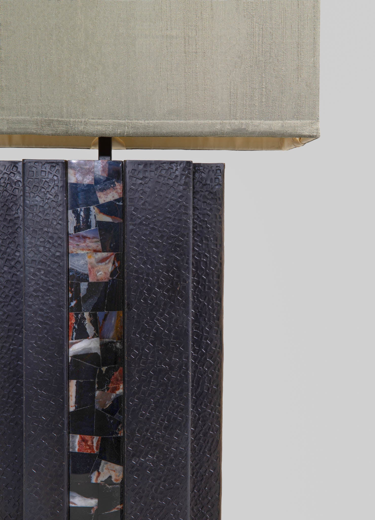 A table lamp with a fabric shade in a neutral beige tone and a base made of textured black material with a colorful mosaic accent in earthy tones and black.