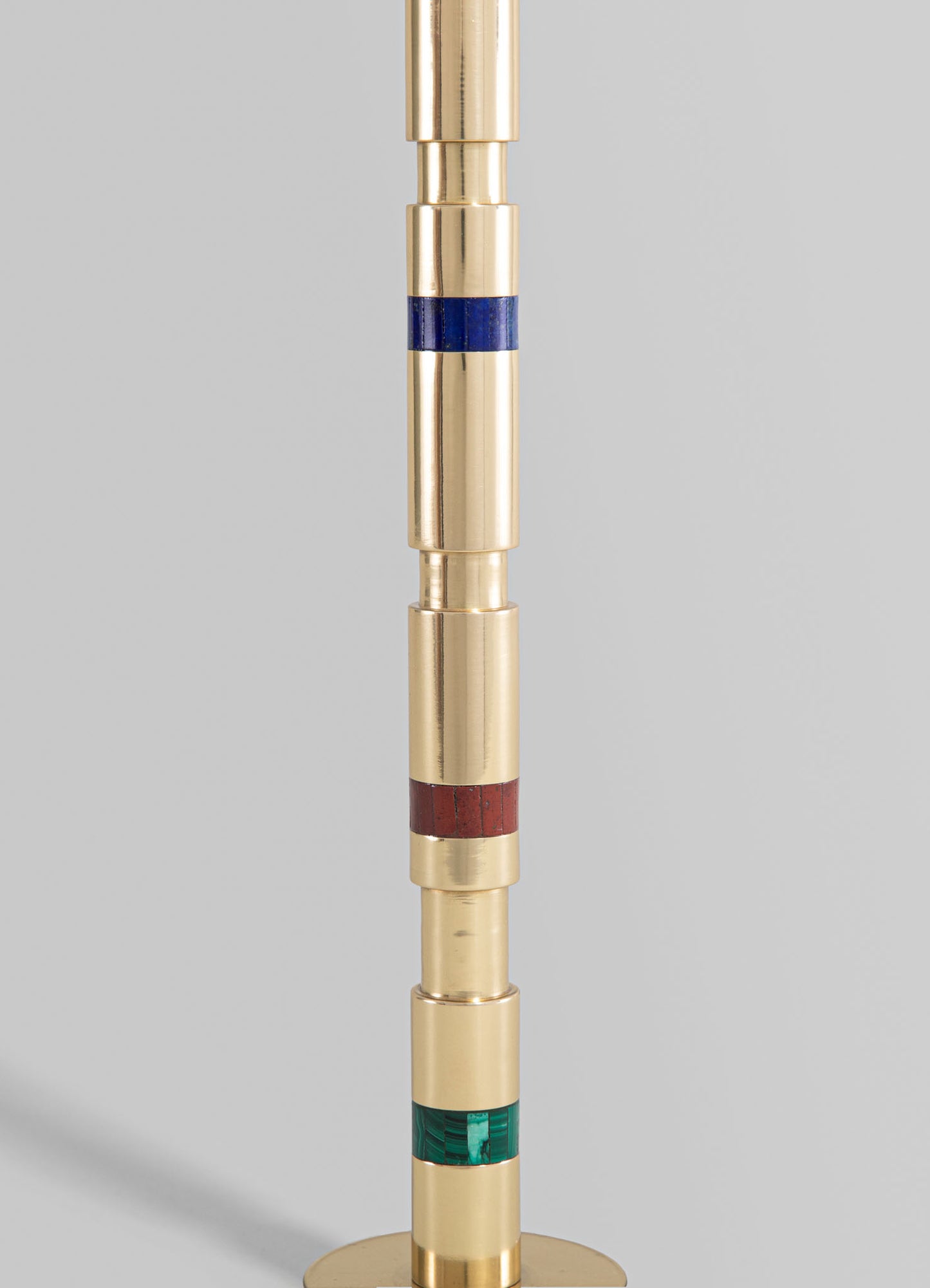 A floor lamp featuring a vertical column with alternating sections of polished brass and colored inlays in blue, red, and green. The design is modern with a sleek, minimalist aesthetic and a combination of metallic and colorful materials.
