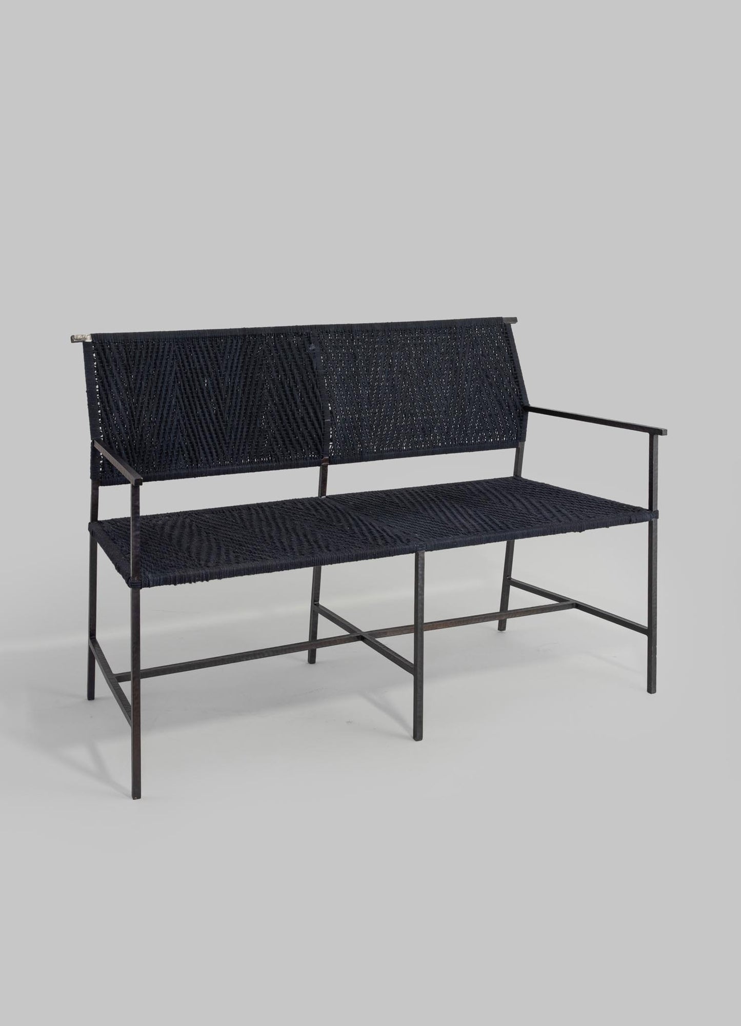 A black metal and woven rattan outdoor bench with a minimalist design, featuring a slatted backrest and armrests for seating.