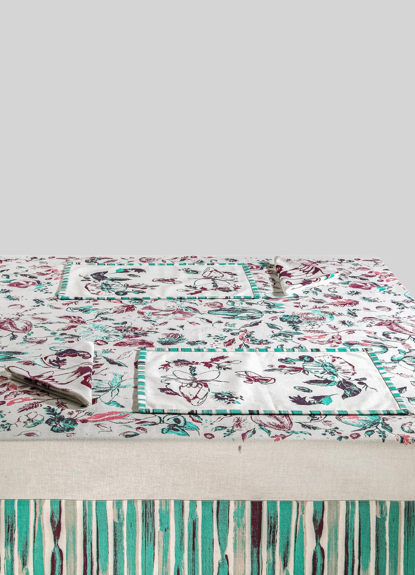 Table linen set with floral and botanical motifs on a white fabric background, including placemats and napkins with a striped border in teal, burgundy, and black tones. The tablecloth features vertical striped pattern in matching colors at the border.
